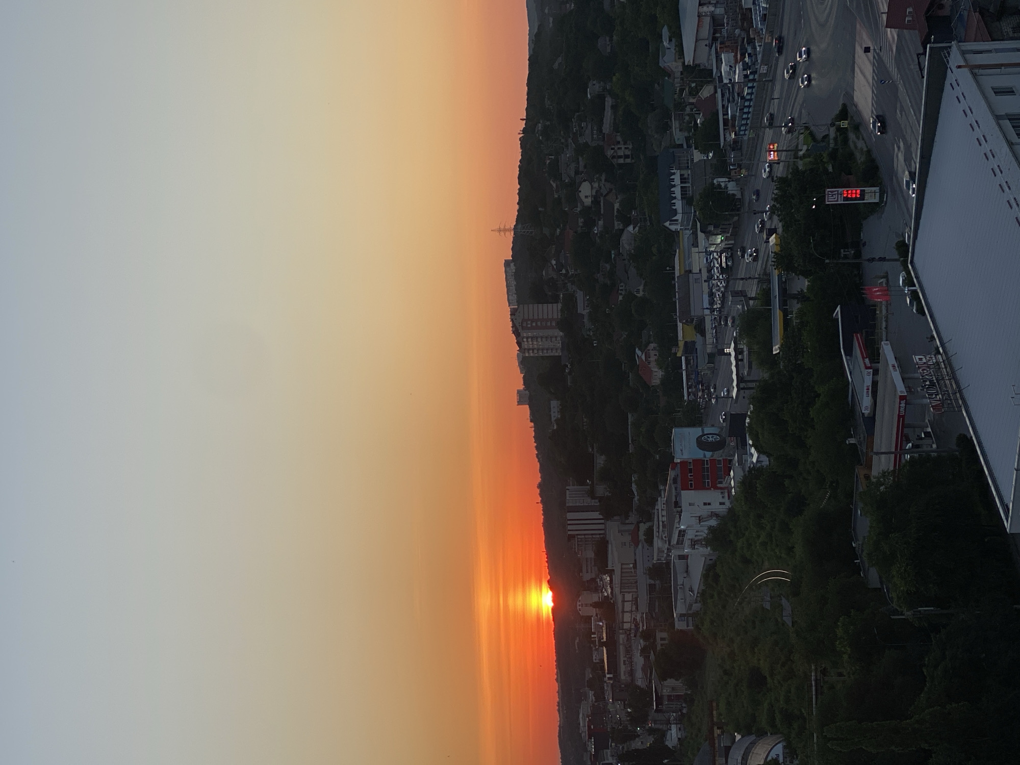 The image shows a sunset over a city that appears to be Chișinău, Moldova. The sun, a bright orange orb, is setting behind a tree-covered hill, casting a warm glow across the sky. The sky transitions from a pale blue at the top to a vibrant orange near the horizon.
In the foreground, there is a cityscape with various buildings, including residential and commercial structures. Some buildings are modern with glass facades, while others appear older. A road with traffic is visible, with cars and trucks moving along it. There are also signs of industrial activity, such as a gas station with a visible "Lukoil" logo.
The terrain is varied, with hills and lower-lying areas. The vegetation appears lush, with trees and greenery dotting the landscape and covering the hills. The overall impression is of a city at dusk, with the natural beauty of the sunset contrasting with the man-made structures of the urban environment. The image appears to be taken from a high vantage point, providing a panoramic view of the city and its surroundings.