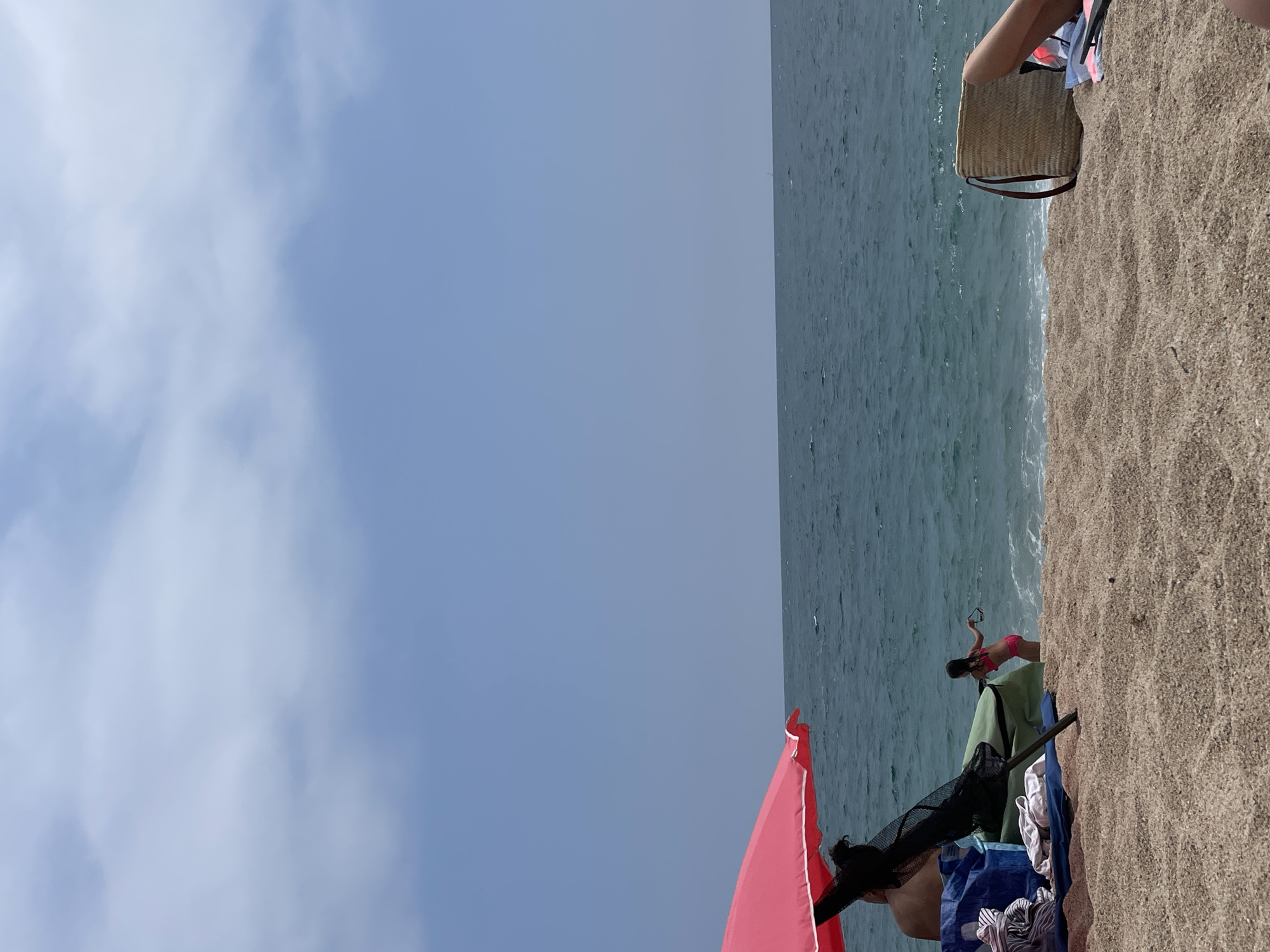 This is a medium shot photograph of a beach scene in Canet de Mar, Spain, taken in daylight. The image is oriented vertically, with the sky occupying the left two-thirds of the frame and the beach and ocean on the right. The sky is a pale blue with scattered white clouds. The ocean is a deep blue-green with gentle waves lapping at the shore. The beach is covered in light brown sand.
In the foreground on the right, a woman in a pink bikini is sitting near some beach items, including a green towel, a black mesh bag, and a blue bag with clothing. Behind her, a red umbrella is partially visible. Further to the right and slightly behind her, a woven straw bag with brown handles rests on the sand. Another person's legs are visible in the upper right corner, suggesting they are sitting on the beach.
The scene appears to be a leisurely day at the beach, with people relaxing by the water. The weather seems pleasant, with a clear sky and calm sea. There is no visible text in the image.