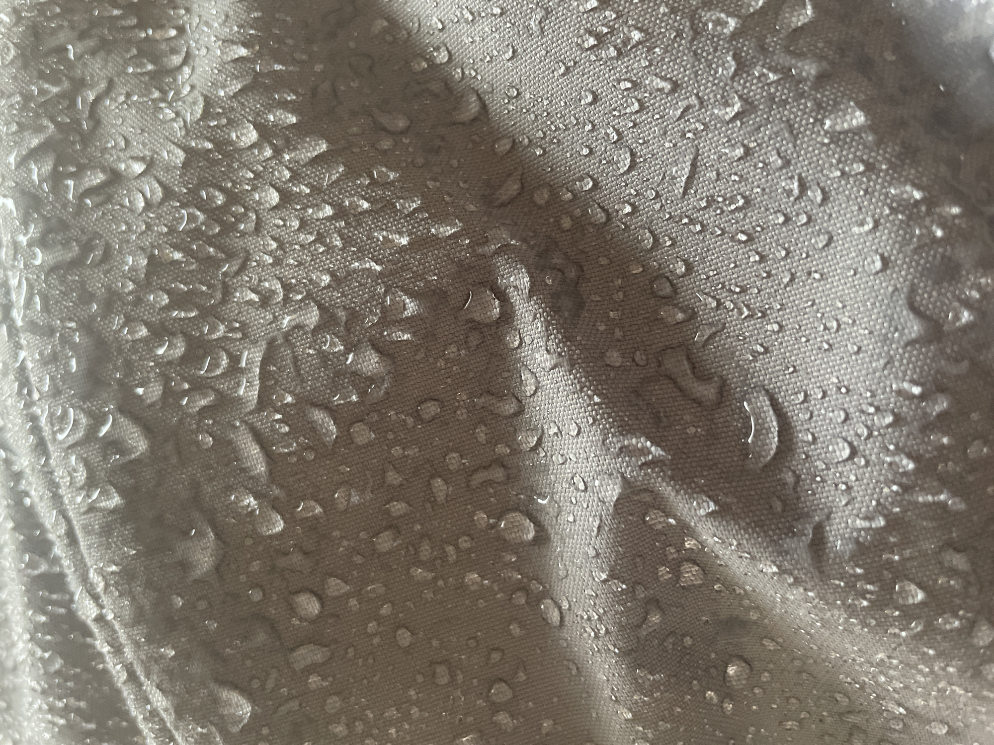 Close-up of a grey fabric surface covered with numerous clear water droplets. The fabric appears to be woven with a visible texture. There are folds and creases in the material, creating variations in light and shadow. The background is out of focus. The image location is unknown and does not contain enough detail to ascertain a specific location in Chaikovsky, Russia, or any other place.