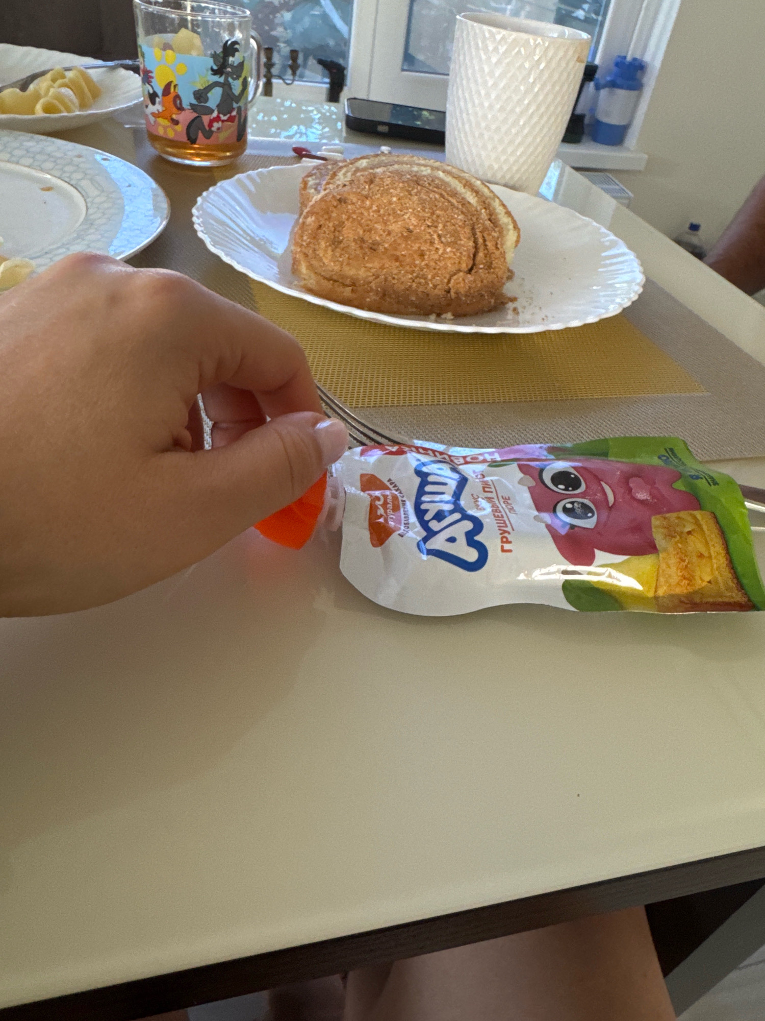 The image depicts a close-up view of a hand holding a fork, which is about to pierce an orange cap on a pouch of fruit puree. The pouch features a cartoon character with large eyes and a pink body. In the background, there are plates with food, a glass with a cartoon image, and a white textured cup. The scene appears to be set on a table, suggesting a mealtime or snack setting. The location is identified as Uyutnoye, Ukraine.