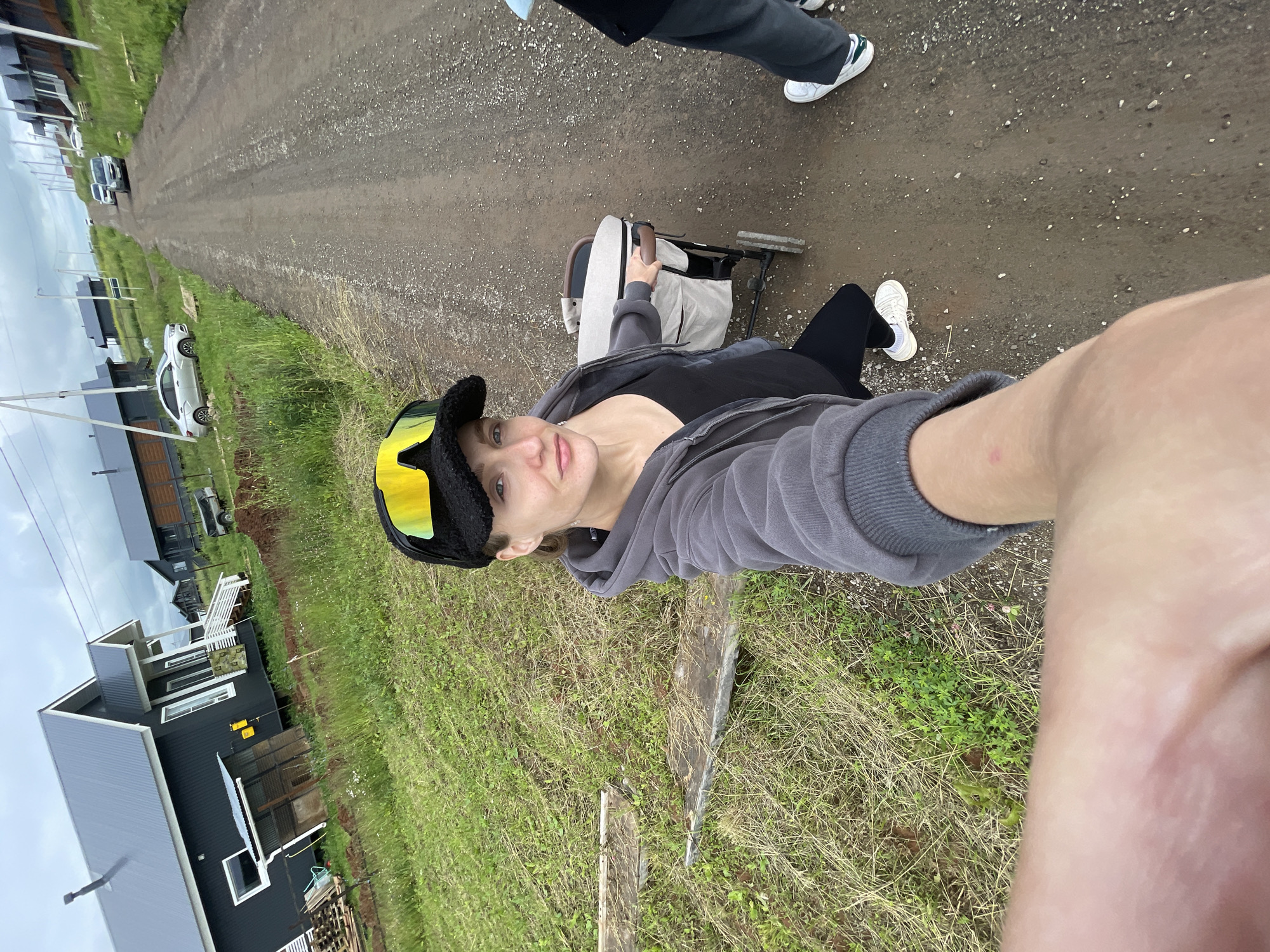 A woman in a grey hooded sweatshirt, black tank top, black leggings, and a black baseball cap with yellow goggles is taking a selfie with a baby stroller. She is standing on a dirt road in what appears to be a rural area, with several modern houses visible on the left side of the road and a line of houses on the right side. The houses are dark-colored with grey roofs. The weather is overcast. A second person, partially visible on the right side of the image, is wearing dark pants and white sneakers. The area is grassy with some areas of exposed dirt. The scene suggests a residential development in a semi-urban or rural setting. The image appears to be taken in the village of Podshivalovo, Russia.