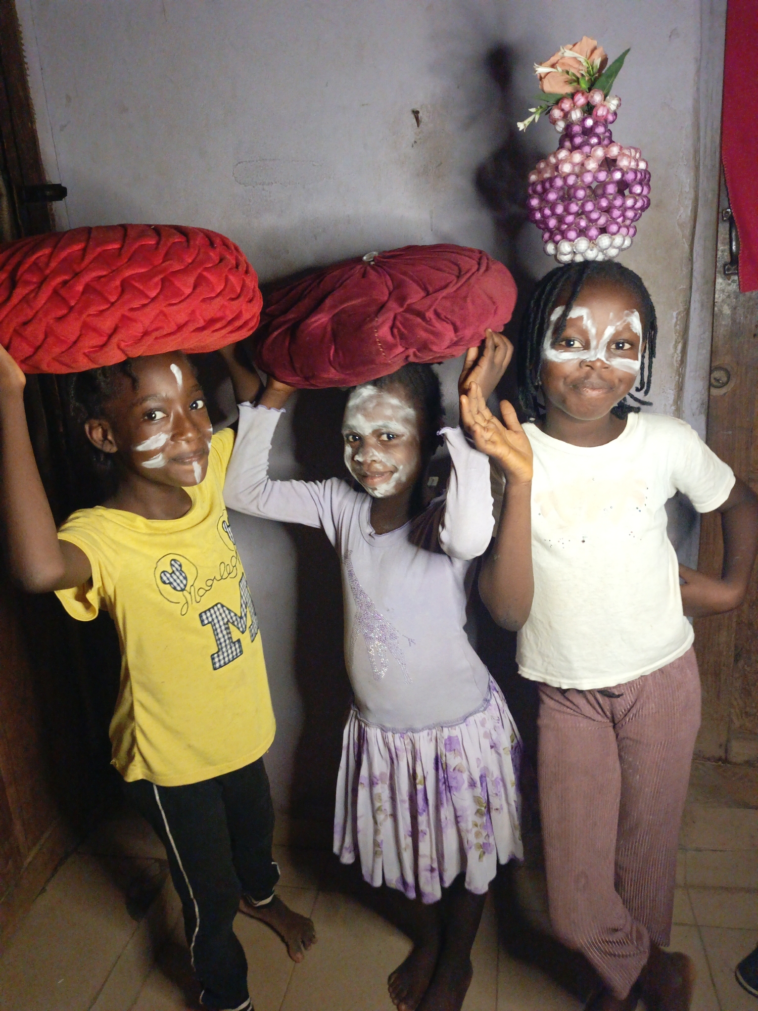 Three young Black girls stand barefoot indoors, posing for the camera with objects balanced on their heads. Each girl has distinctive white face paint applied in patterns across their forehead, nose, and cheeks, suggesting a playful or cultural adornment.
The girl on the left, wearing a bright yellow t-shirt with a Mickey Mouse-like design and the partially visible text "Marley," balances a large, bright red, textured cushion-like object on her head. Her arms are raised to support it.
The middle girl, dressed in a light purple long-sleeved top with glitter details and a floral-patterned skirt, has a dark red or maroon soft, cushion-like object on her head. She also holds her hands up to steady the object.
The girl on the right, in a white t-shirt and brown or mauve ribbed pants, balances a decorative vase on her head. The vase is crafted from purple, pink, and white beads and contains artificial pink and white flowers. She smiles directly at the camera.
The background consists of a light gray or off-white wall, with a wooden doorframe visible on either side. The floor appears to be a light-colored tile or concrete. The indoor lighting is even, suggesting an artificial source or diffused natural light. All three girls are smiling, conveying a sense of joy and participation in an activity.
