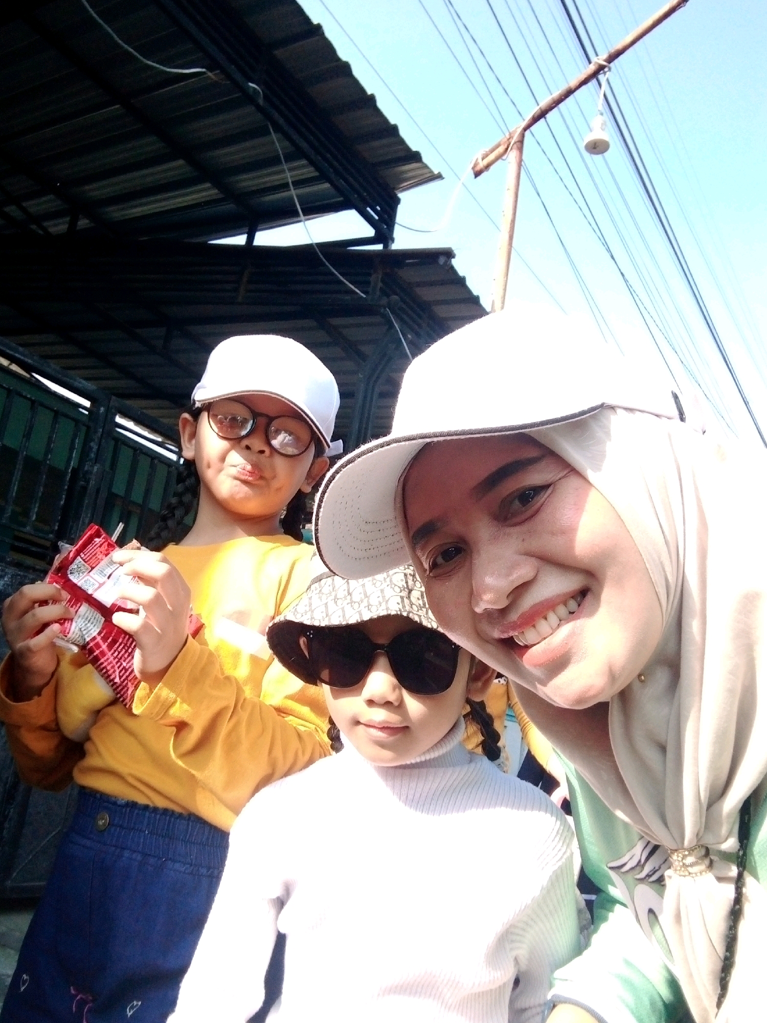 A selfie shows a woman and two children outdoors in what appears to be a residential area. The woman, wearing a light-colored hijab and a white cap, is smiling widely. To her left, a child in a white turtleneck, sunglasses, and a patterned bucket hat faces forward. Further left, another child wearing a yellow long-sleeved shirt, glasses, and a white cap holds a snack package. The background reveals a corrugated metal roof, some greenery, and a blue sky with electrical wires. The image quality is slightly blurred, suggesting it was taken with a mobile phone.