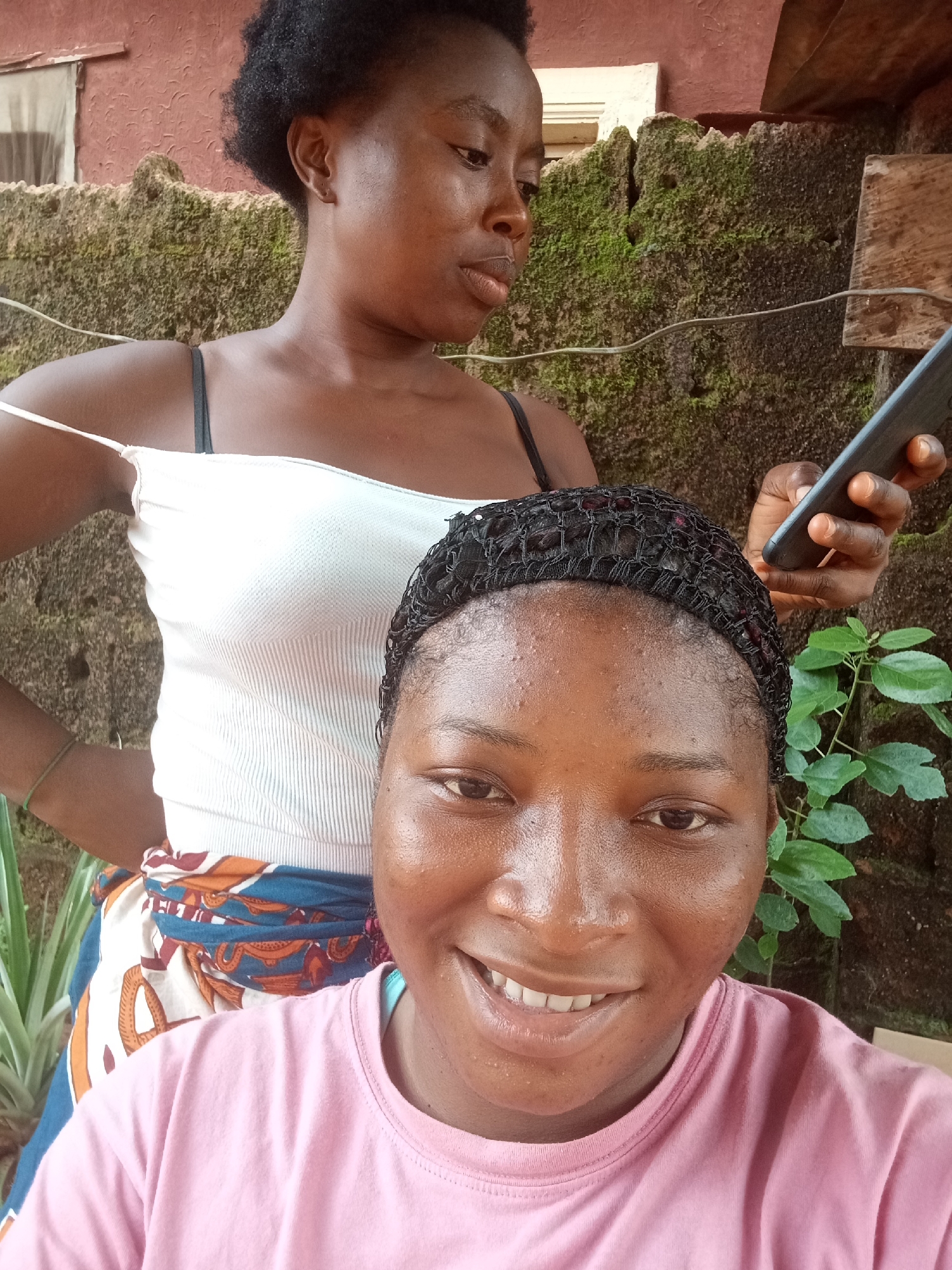 Two women are in the image. One, in the foreground, wears a pink shirt and smiles at the camera. Her hair is covered with a black netting. Behind her is another woman wearing a white top with black straps. She looks to the side while holding a cell phone. The woman in the white top has a wrap tied around her waist. They appear to be outdoors, against a wall covered with moss and some greenery. A window can be seen above them. The city of Ogidi, Nigeria, cannot be confirmed definitively from this image, though the subjects are likely of Nigerian descent given their features and the style of dress, e.g. the wrap tied around the waist.
