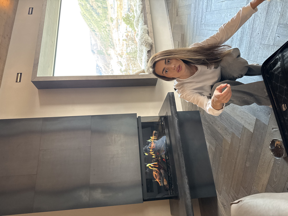 An indoor scene in Schladming, Austria, displays a modern living space overlooking a snow-dusted mountainous landscape. In the immediate foreground, a female subject with long, light brown hair, wearing a white long-sleeved shirt and grey pants, crouches on a herringbone-patterned wooden floor. Her right arm is extended forward with an open hand, while her left hand is near a partially open black bag with a quilted pattern. To the bottom left of the frame, a small metallic bowl containing dark pellet-like objects is visible. To the female subject's left, a contemporary fireplace features a tall, dark, multi-paneled facade, with a glass-enclosed firebox containing artificial logs and active flames. A dark, wide hearth extends from the base of the fireplace. To the subject's right and further back, a large, dark-framed window reveals an exterior view of snow-covered mountains, evergreen trees, and scattered buildings in a valley. A wide window seat covered with white, shaggy material is situated beneath the window. The interior walls are a light, neutral color, and recessed light fixtures are visible on the wall above the window.