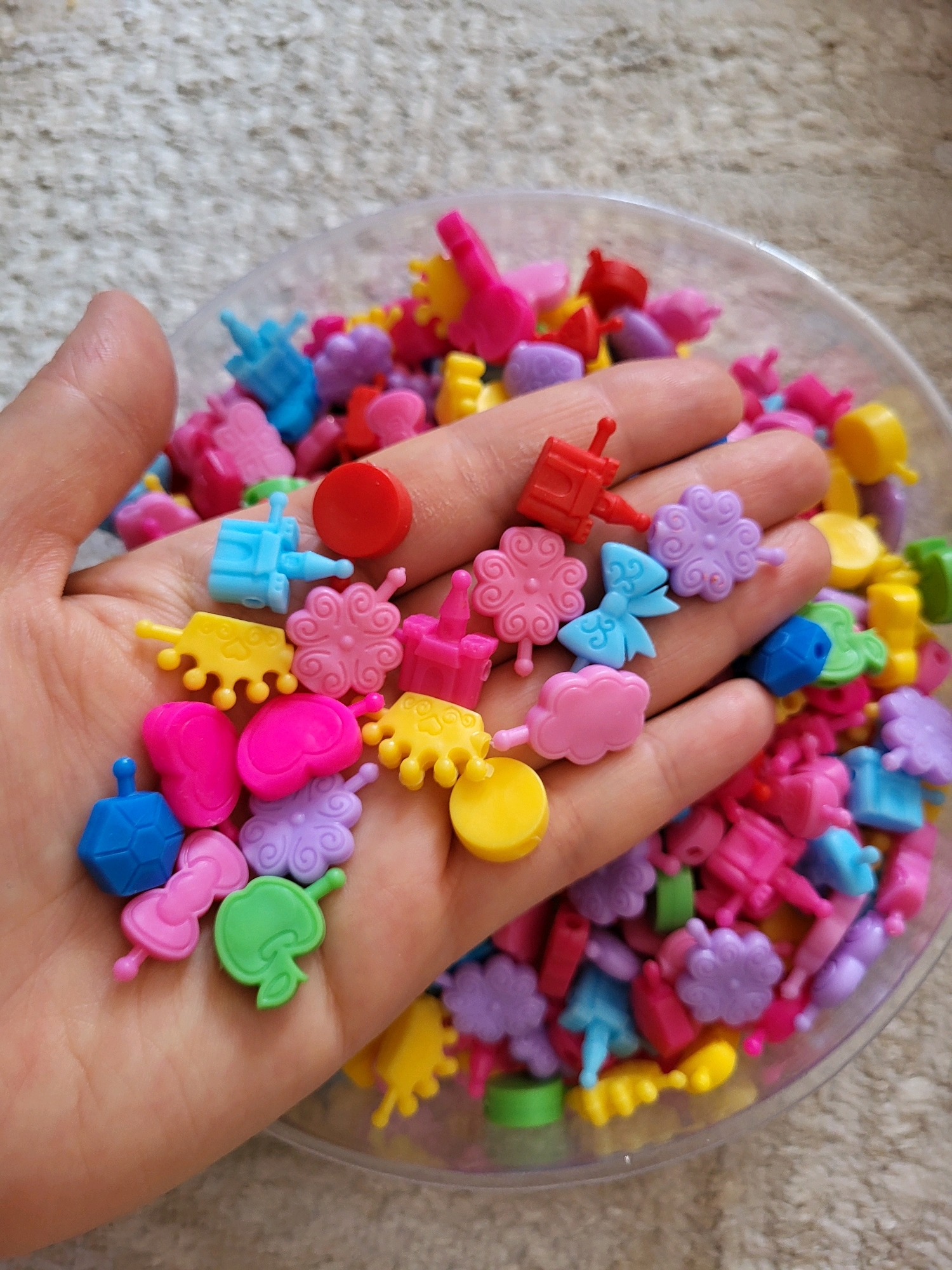 A close-up, high-angle shot shows a person's open palm, which is filled with numerous small, brightly colored plastic charms. The charms are diverse in shape and color, including hearts, bows, flowers, clouds, crowns, and abstract geometric shapes, in shades of pink, blue, yellow, purple, red, and green. The hand is positioned over a clear plastic container that holds a larger quantity of similar charms, some of which are visible at the bottom right of the frame. The background is a textured, light-colored surface, likely a carpet or woven fabric. The scene appears to be indoors, with the lighting suggesting natural daylight. There is no indication of the city of Almaty, Kazakhstan, or any urban environment in the image.