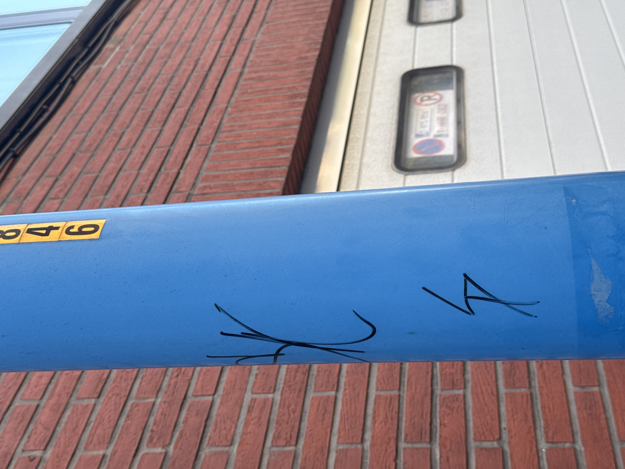 In Strombeek-Bever, Belgium, a bright blue cylindrical pole stands in front of a red brick building and a white garage door. A yellow sticker with the numbers "846" is affixed to the upper portion of the pole. Black graffiti marks are present on the pole, with a complex symbol resembling an abstract "X" or intertwining lines positioned in the middle, and a lightning bolt-like symbol further down. The garage door has two rectangular windows, one of which contains signage with a red circle and diagonal line crossed with a blue circle and red R, indicating parking restrictions. The environment suggests an outdoor urban or commercial setting.