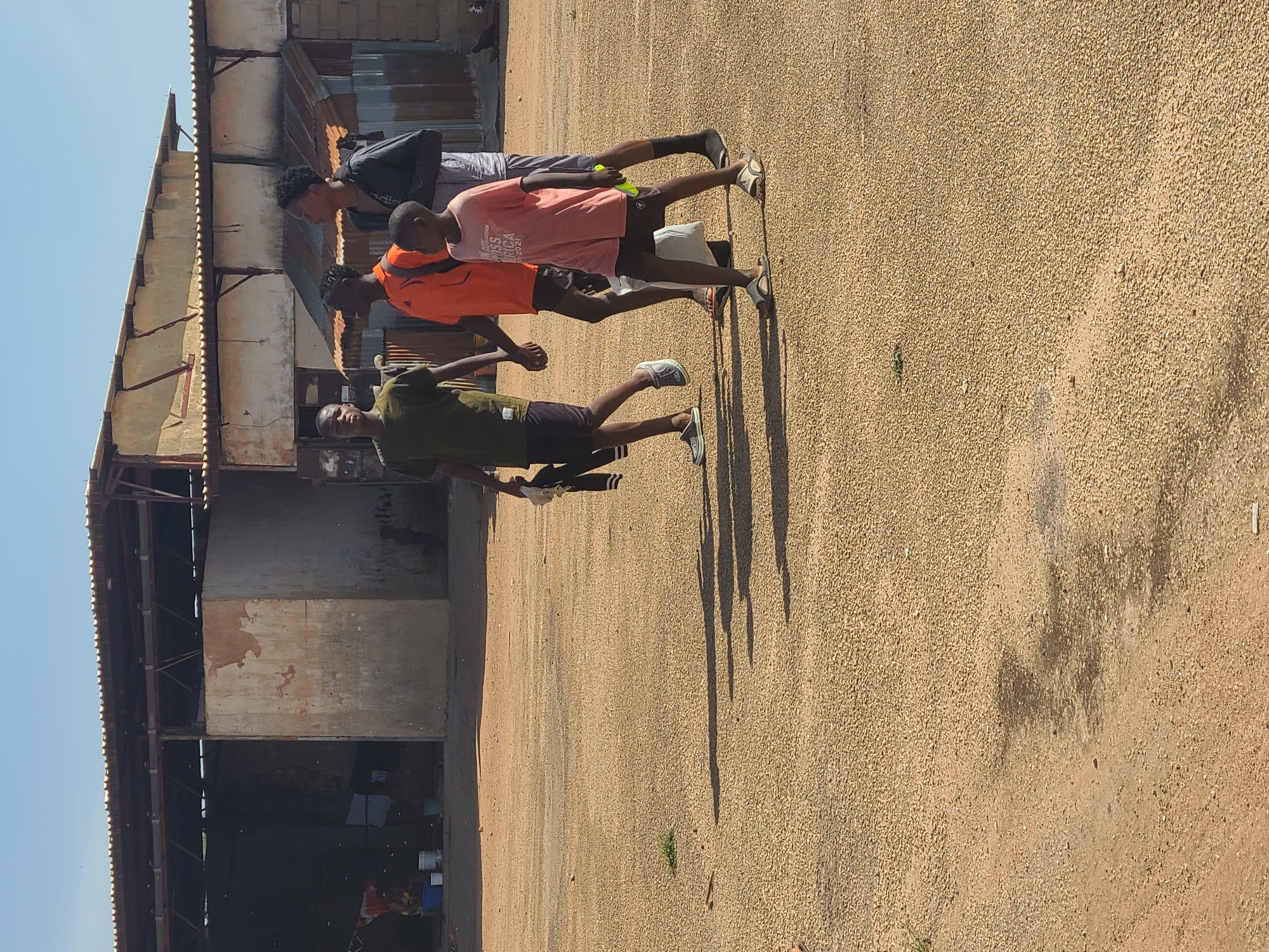 The image displays five young men standing outdoors on a bright, sunny day in what appears to be Jos, Nigeria. They are positioned on a light brown, gravelly ground surface. In the background, there is a simple, multi-story building featuring a weathered corrugated metal roof and exposed wooden beams. The building's walls are a mix of concrete and exposed brickwork, with the lower level having open sections revealing some stored items, including clothes and buckets.
From left to right, the closest man wears a dark green t-shirt and dark shorts, holding a white object (possibly a phone) and a small, bright yellow-green item. Next to him, another man is dressed in an orange t-shirt and dark shorts. Further back, a third man wears a light pink/peach t-shirt with the text "MISS AFRICA 2021" visible on the front; he also holds a small yellow-green object similar to the first man's. Behind him, a fourth man is in a black jacket, possibly with a partial 'adidas' logo on the sleeve, and grey shorts. A fifth person is partially visible behind the man in the pink shirt.
The strong, defined shadows cast by the individuals on the ground indicate that it is mid-morning or early afternoon under clear, sunny skies. The men appear to be gathered casually, possibly observing something or taking a break. The yellow-green objects in their hands might suggest involvement in a game or activity, though its precise nature is unclear. The overall setting reflects a common architectural style and environment found in the region.