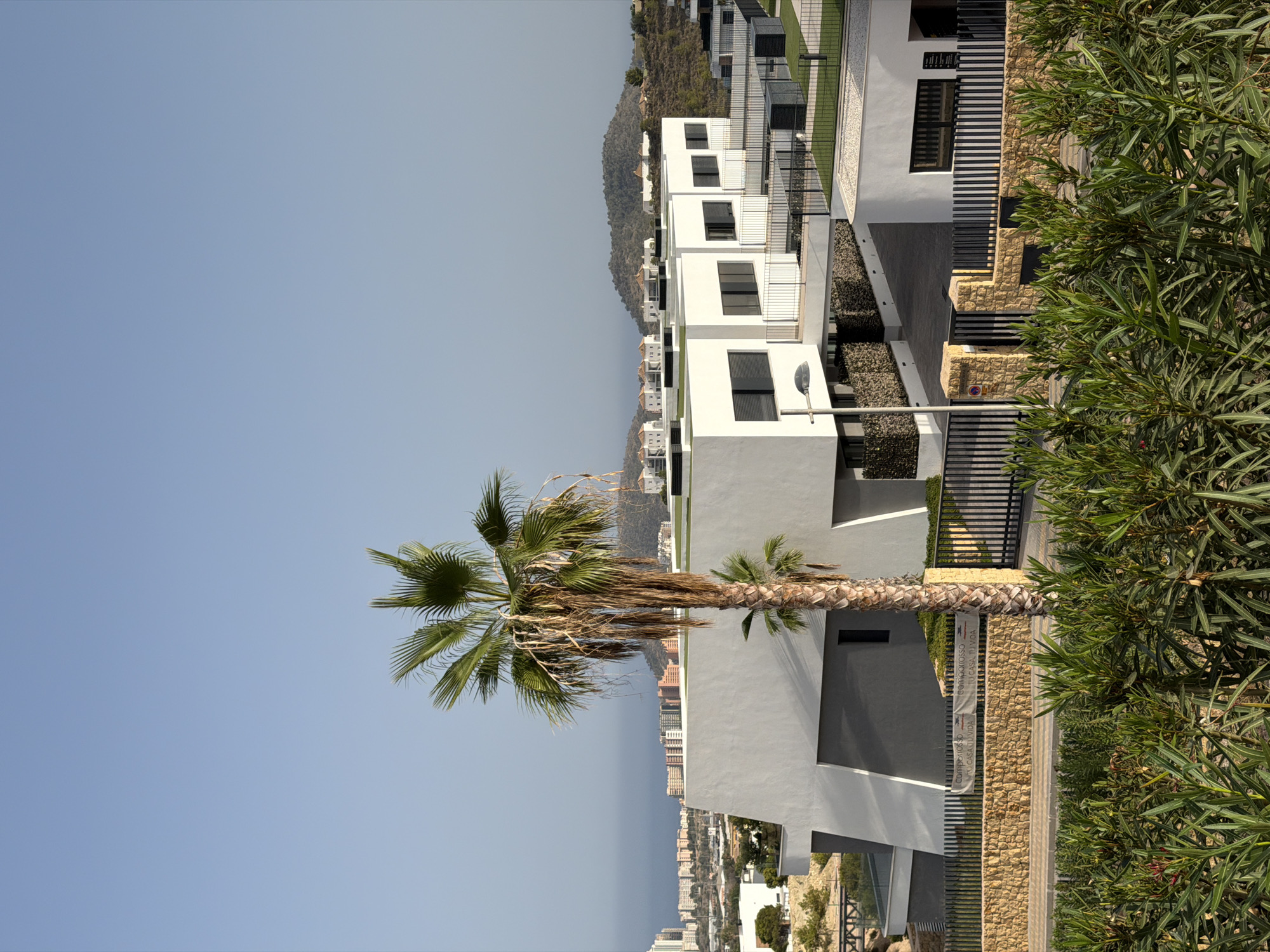 The photograph showcases a modern residential complex in Finestrat, Spain, under a clear, pale blue sky. In the foreground, lush green foliage partially obscures the view of the property. A tall palm tree with dry, drooping fronds stands prominently to the left of the frame, with a smaller palm at its base.
The architecture features stark white, geometric buildings with dark-framed windows. The buildings are constructed with clean lines and flat roofs, some of which are topped with green, artificial turf. A significant structure on the left has a dynamic, angular design with contrasting grey and white sections. A stone wall, constructed from rough-hewn blocks, runs horizontally across the middle ground, punctuated by black metal fencing. A gate is visible within this fencing, and a banner with Spanish text, possibly a real estate advertisement, is affixed to the wall.
In the background, a distant cityscape with numerous buildings, likely apartments or hotels, is visible along the coastline, extending towards the horizon. Rolling, tree-covered hills form the backdrop, with a few scattered white buildings perched on their slopes. The overall impression is one of contemporary design set against a Mediterranean landscape. There are no people visible in the image.