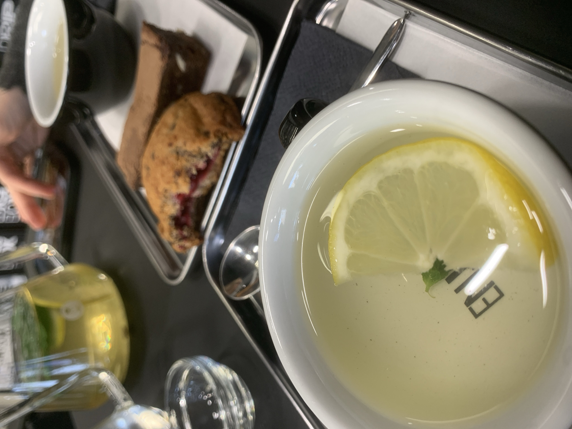 The image shows a tray with refreshments. A cup of lemon tea, a baked treat (likely a muffin with visible fruit filling), and a slice of cake or brownie are presented. There's also a glass jar and another black cup that appears to contain a beverage. The setting is likely a cafe or restaurant. The city or specific details about Izhevsk, Russia, are not evident in this image.