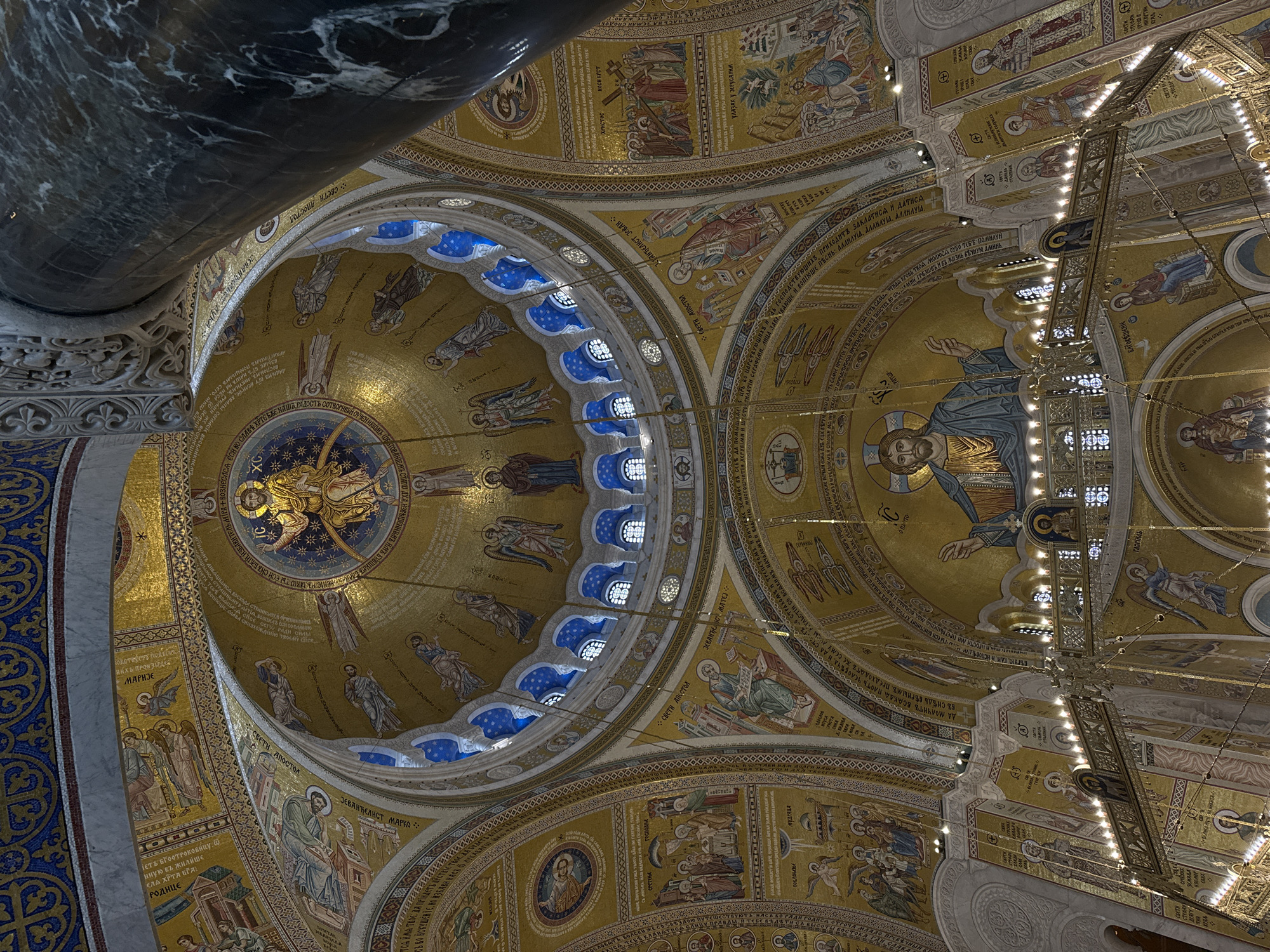 The image captures a portion of the interior dome mosaics of the Church of Saint Sava in Belgrade, Serbia. The shot is taken from below, looking upwards.
Visible are several sections of ornate mosaic artwork featuring depictions of religious figures and biblical scenes rendered in gold, blue, and other colors. The mosaics are arranged in concentric circles and patterns, common in Orthodox church architecture. There is an ornate chandelier and the dark marble column. The presence of Cyrillic text in the mosaics provides further evidence for the Serbian Orthodox context.
The Church of Saint Sava is a prominent landmark in Belgrade, known for its grandeur and Byzantine-style mosaics, the image offers a view of the church's detailed interior decor.
