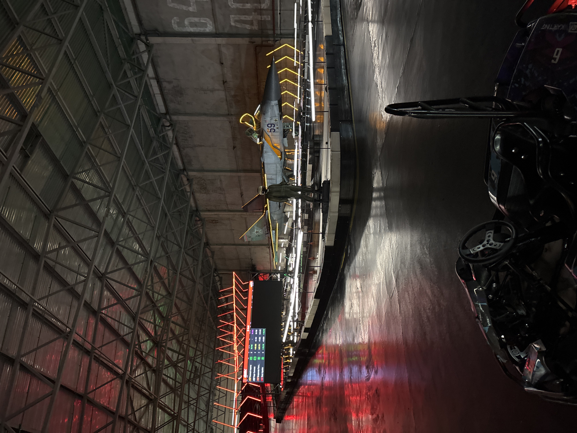 The image depicts the interior of an indoor go-kart track located in Saint Petersburg, Russia, at the karting complex "Pitstop". The scene is illuminated by vibrant red and yellow neon lights, reflecting off the wet asphalt floor. In the background, a model airplane, identified as a MiG-21 with the number "59", is prominently displayed, surrounded by yellow lighting. Adjacent to the airplane is a life-sized mannequin dressed in a pilot's uniform. A large digital scoreboard displaying race times and driver information is positioned on the left side of the track, with red neon lights forming an abstract design above it. In the foreground, the steering wheel and cockpit of a go-kart are visible, suggesting the presence of active participants or ready-to-use karts. The overall environment appears to be a hangar or large industrial space, with a corrugated metal roof supported by a steel truss system. The walls are concrete, with large numbers "64" and "40" painted on them. The lighting creates a dramatic and energetic atmosphere.