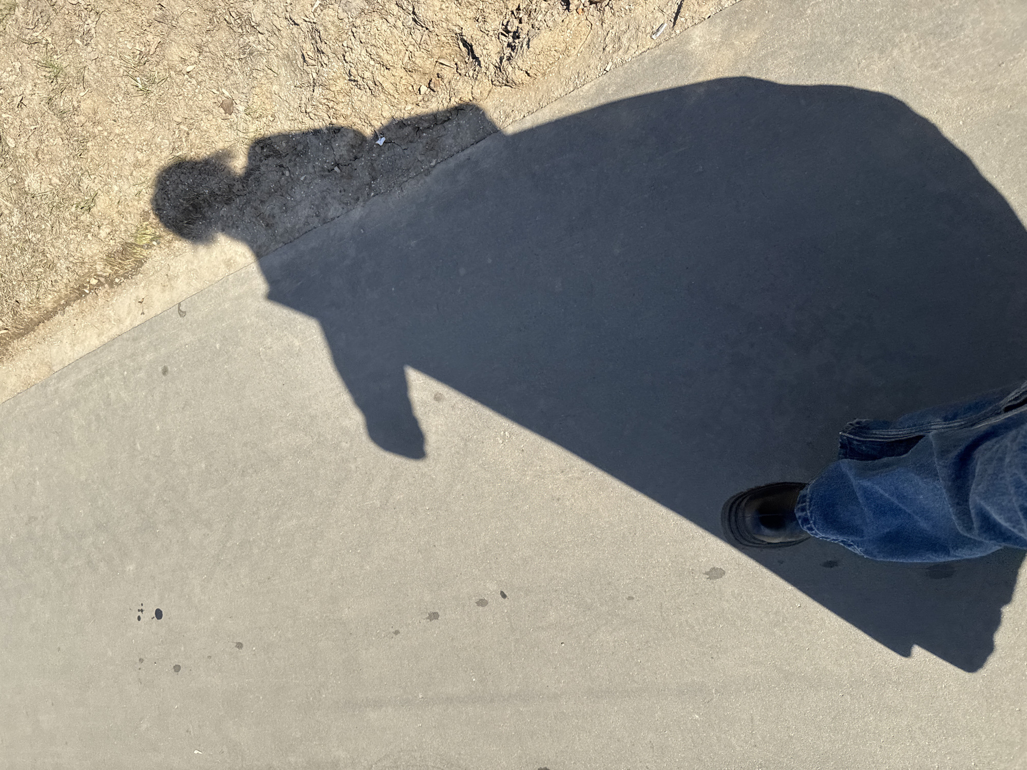 The image shows a shadow of a person and a foot with a shoe on a paved surface, possibly a sidewalk. The shadow suggests the person is wearing a wide-brimmed hat or has a large head covering. The foot is clad in a black shoe and blue denim pants. There are some dark spots on the pavement. A patch of dirt and vegetation is visible in the background. No indication of the city Khimki, Russia is visible.