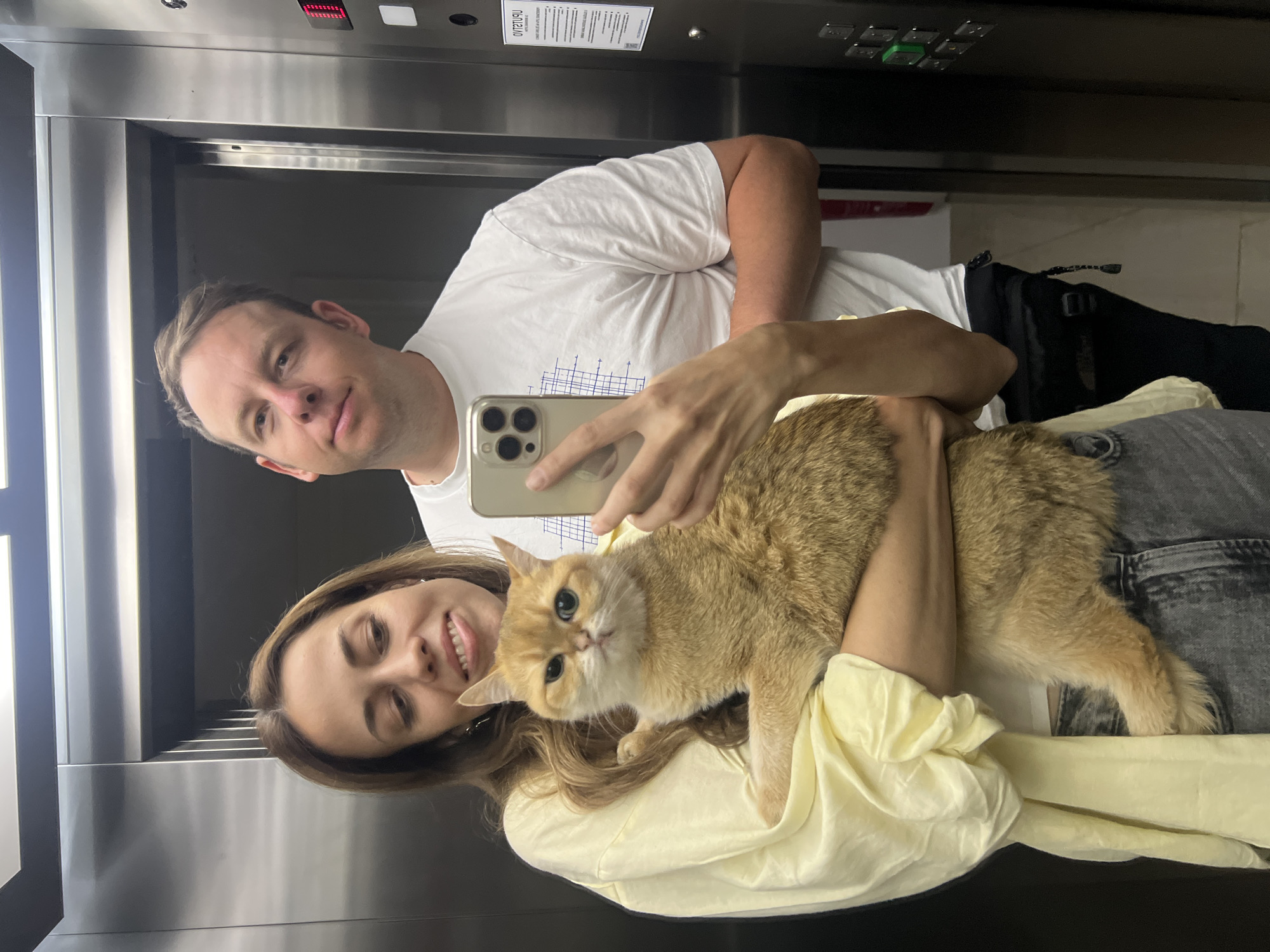 A couple takes a selfie with their cat in an elevator. The man is on the left, wearing a white t-shirt, and holds the phone. The woman is on the right, wearing a yellow shirt, holding a golden cat. The elevator has stainless steel walls and control panels. The location is Bar, Montenegro, based on the surroundings.