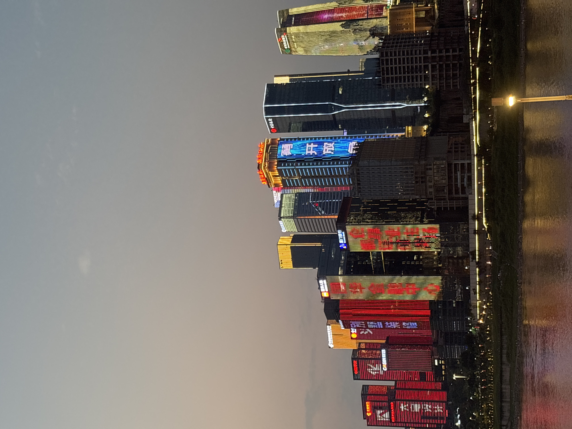 The image shows a nighttime view of buildings in the Yuzhong District of Chongqing, China. The buildings are tall and brightly lit with neon lights in various colors, including red, blue, and yellow. The lights reflect off the water in the river that runs along the edge of the buildings. There are street lights and vegetation visible along the riverbank. The sky is dark, suggesting it is evening or night. The scene appears to be a modern urban landscape with a focus on architectural lighting and a waterside location.