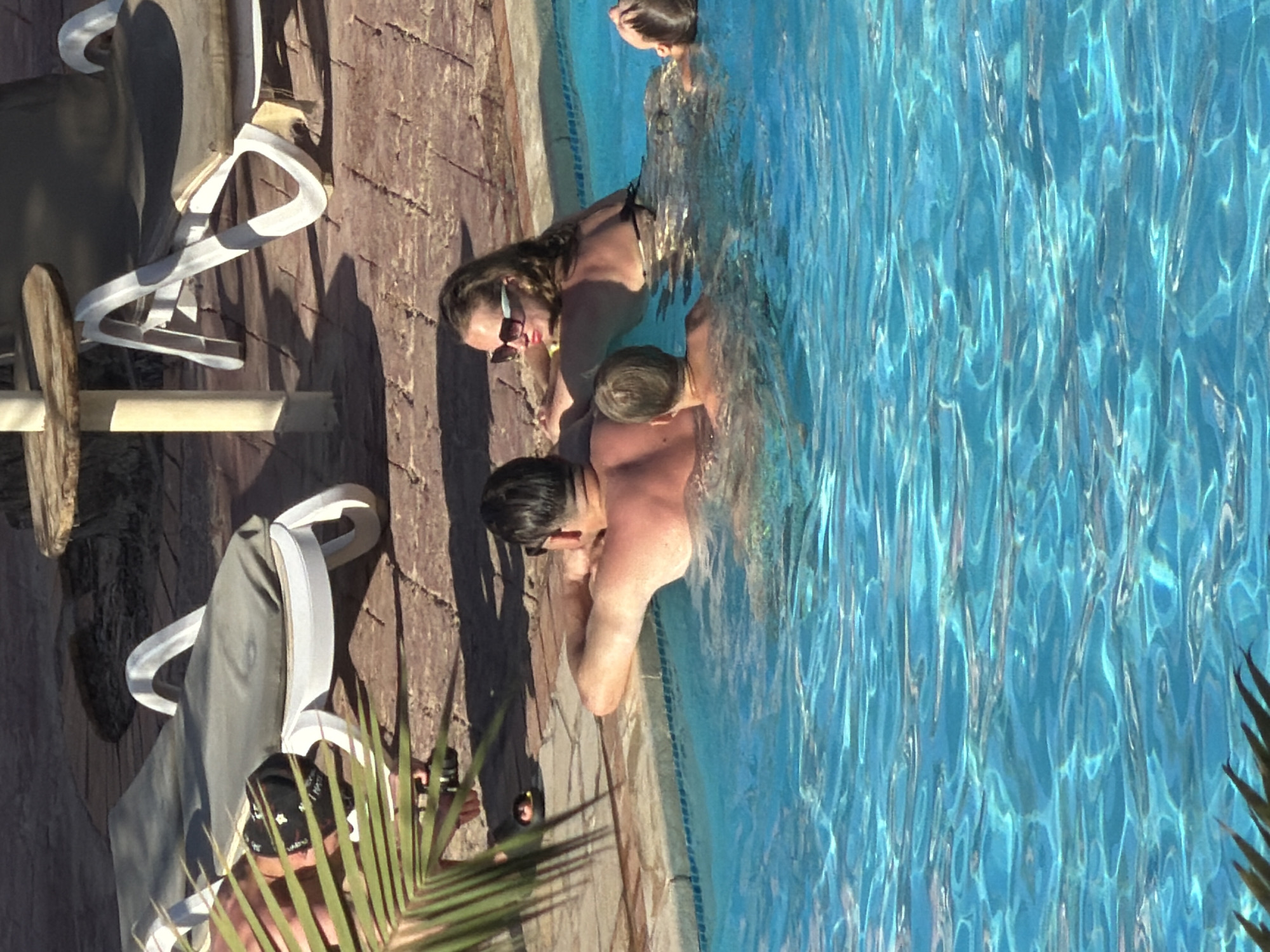 In Sharm El Sheikh, Egypt, a family is enjoying a swimming pool. Two adults, a man and a woman, are partially submerged in the water, their backs to the camera. The man is wearing dark sunglasses. The woman is wearing a bikini and sunglasses, her head resting on her arm. A young boy is also in the pool, facing away from the camera. Another child, a girl, is visible to the right of the woman, looking upwards. In the background, two white lounge chairs are positioned on a brick patio, and a small wooden table is nearby. A palm frond extends into the lower-left corner of the frame, casting a shadow. The water in the pool is bright blue and reflects sunlight.
