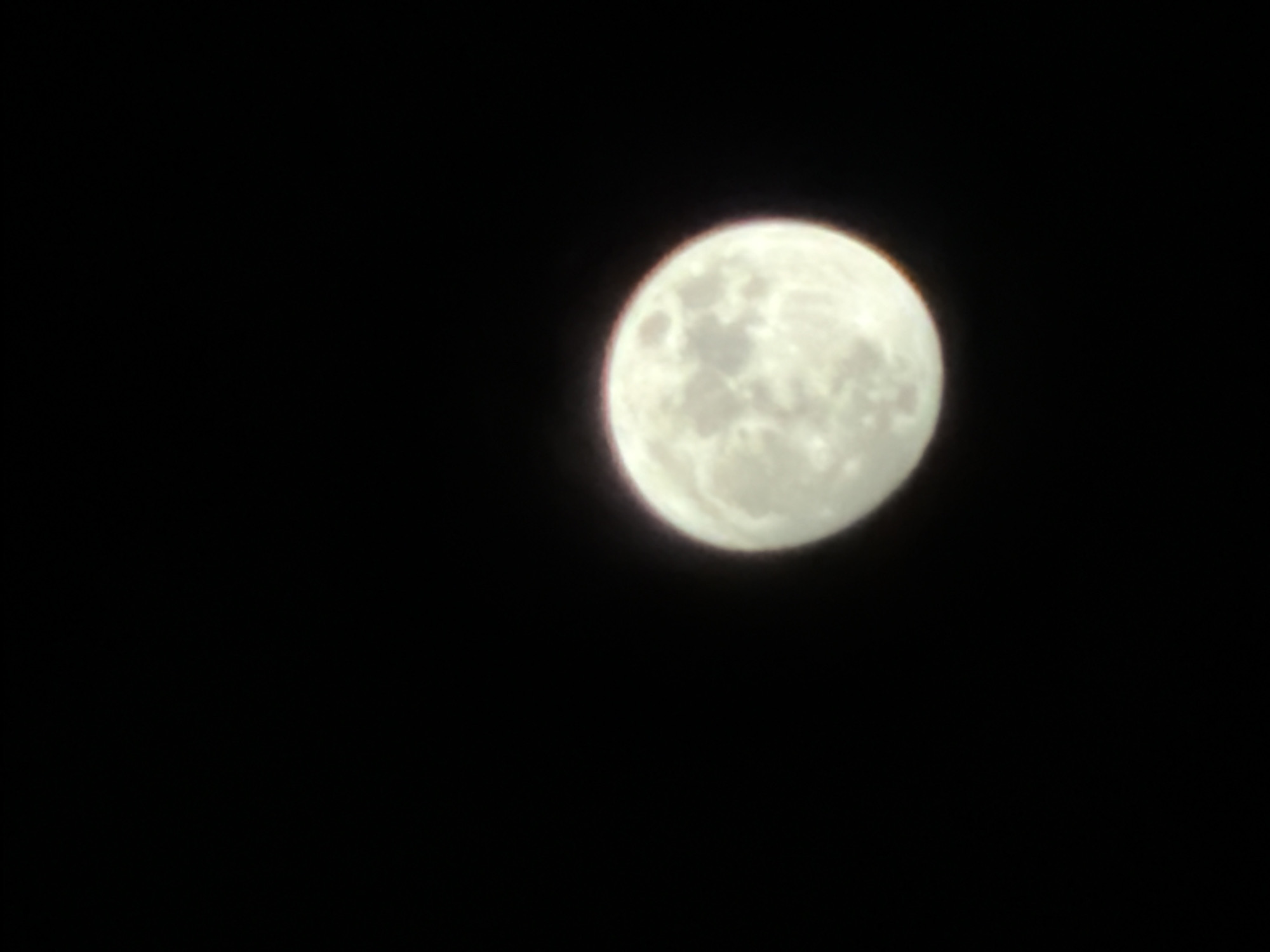 The media file contains a close-up, slightly out-of-focus shot of the moon against a black sky. The moon is full and appears bright white with visible craters and surface details. It occupies the right side of the frame, leaving a large expanse of dark space to the left. There is a subtle reddish-orange halo around the upper edge of the moon, possibly due to atmospheric conditions or lens effects. The image is dark, suggesting it was taken at night. No people, text, or specific location cues are visible.