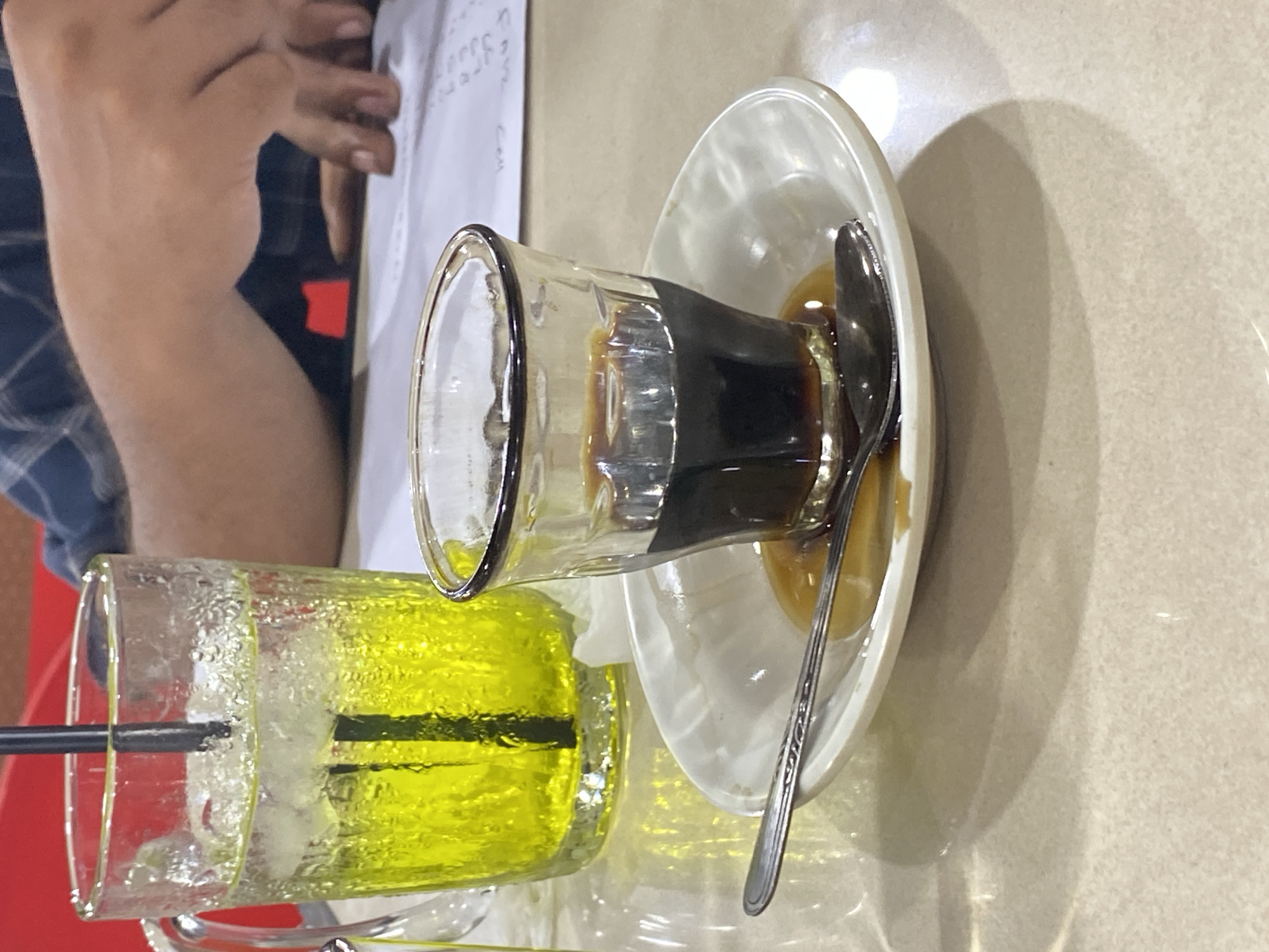 In the city of Lhokseumawe, Indonesia, on a light-colored, textured table, a glass mug filled with a bright yellow, ice-cold beverage and a black straw stands to the left. In the foreground, a small clear glass containing a dark liquid, likely coffee, sits on a white saucer. A silver spoon rests on the saucer, partly submerged in a small amount of spilled liquid. In the background, a person's hands are visible, resting on a white surface with faint text, suggesting a document or paper. The overall scene depicts a casual moment, possibly at a cafe or home, with two drinks and a person engaged with some paperwork.