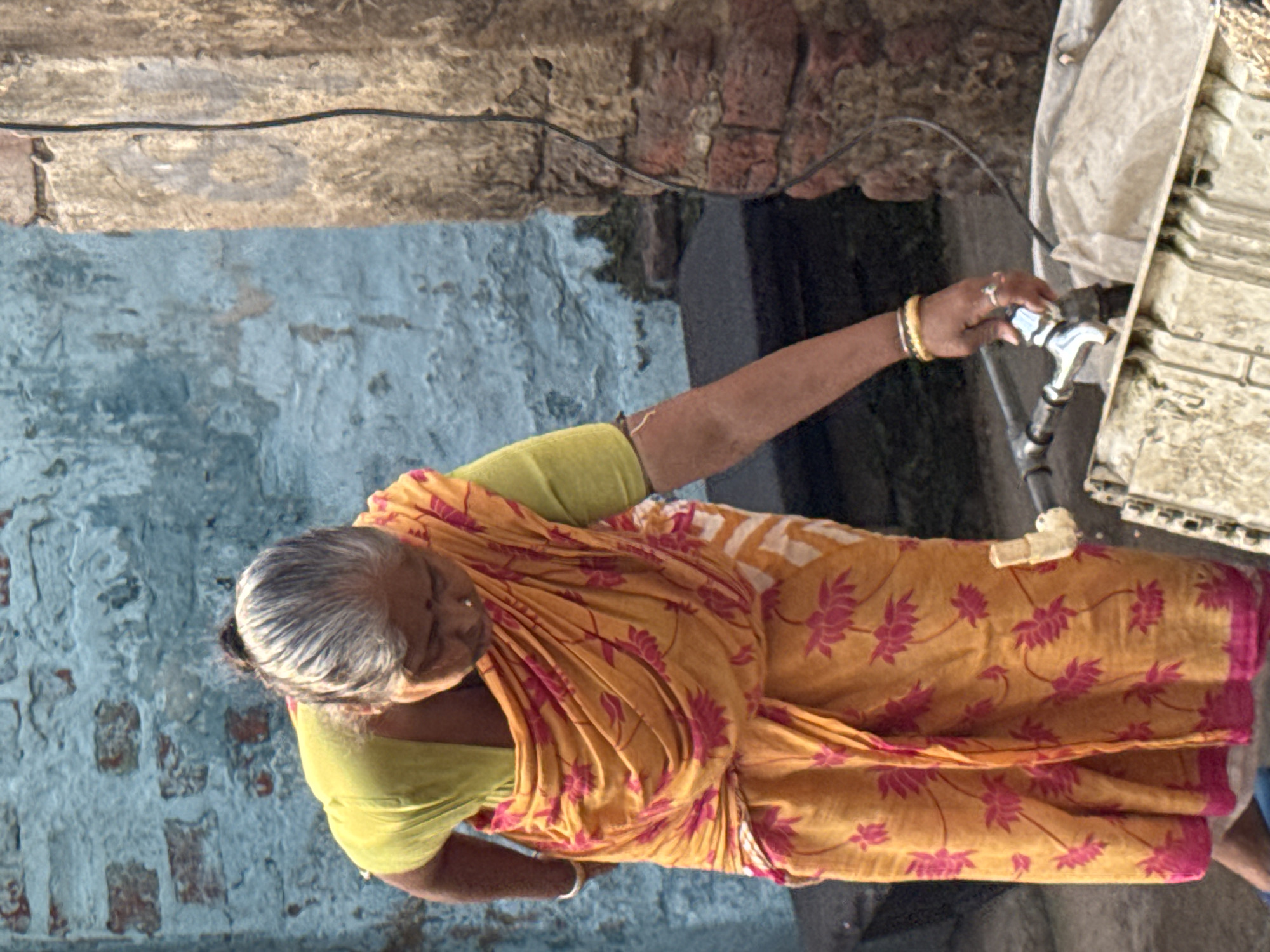 An older woman with short, gray hair, wearing a yellow sari with pink floral prints, reaches out to turn on a silver faucet. The background consists of a light blue, rough brick wall and a gray wall with a black wire running across it. A white, boxy object is visible to the right of the faucet. The location is Kolkata, India.