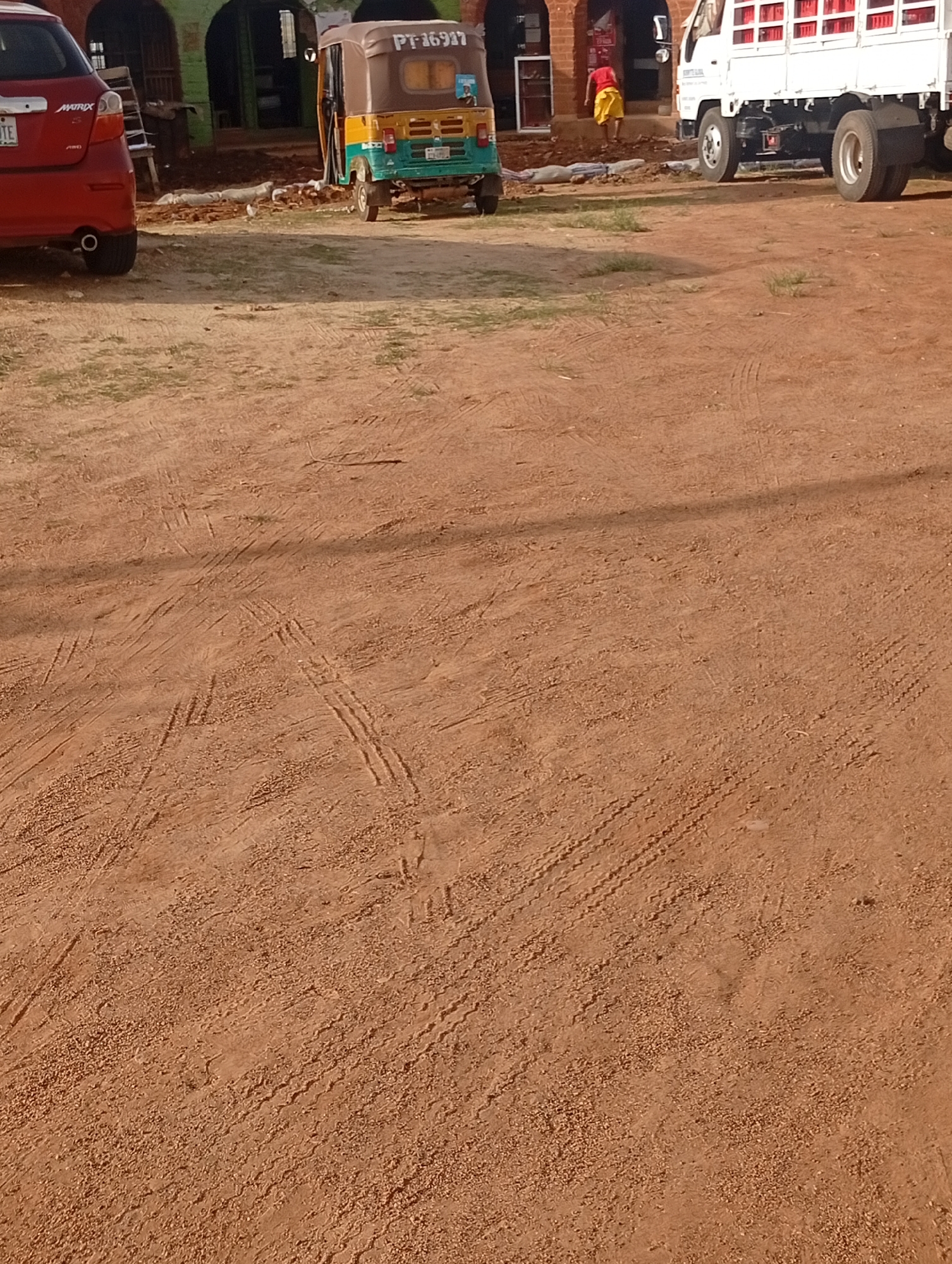 In a dusty, outdoor environment, likely in Jos, Nigeria, a red Toyota Matrix car is parked on the left. To its right, a three-wheeled auto-rickshaw, identified by the license plate "PT 16917," is positioned. Further to the right, a white truck with red crates loaded on its bed is visible. A person in a yellow skirt and red shirt is walking away from the white truck and into a building with an arched entrance. The ground is composed of reddish-brown dirt with visible tire tracks from various vehicles. The overall scene suggests a commercial or transportation hub in a dry, possibly arid, region.