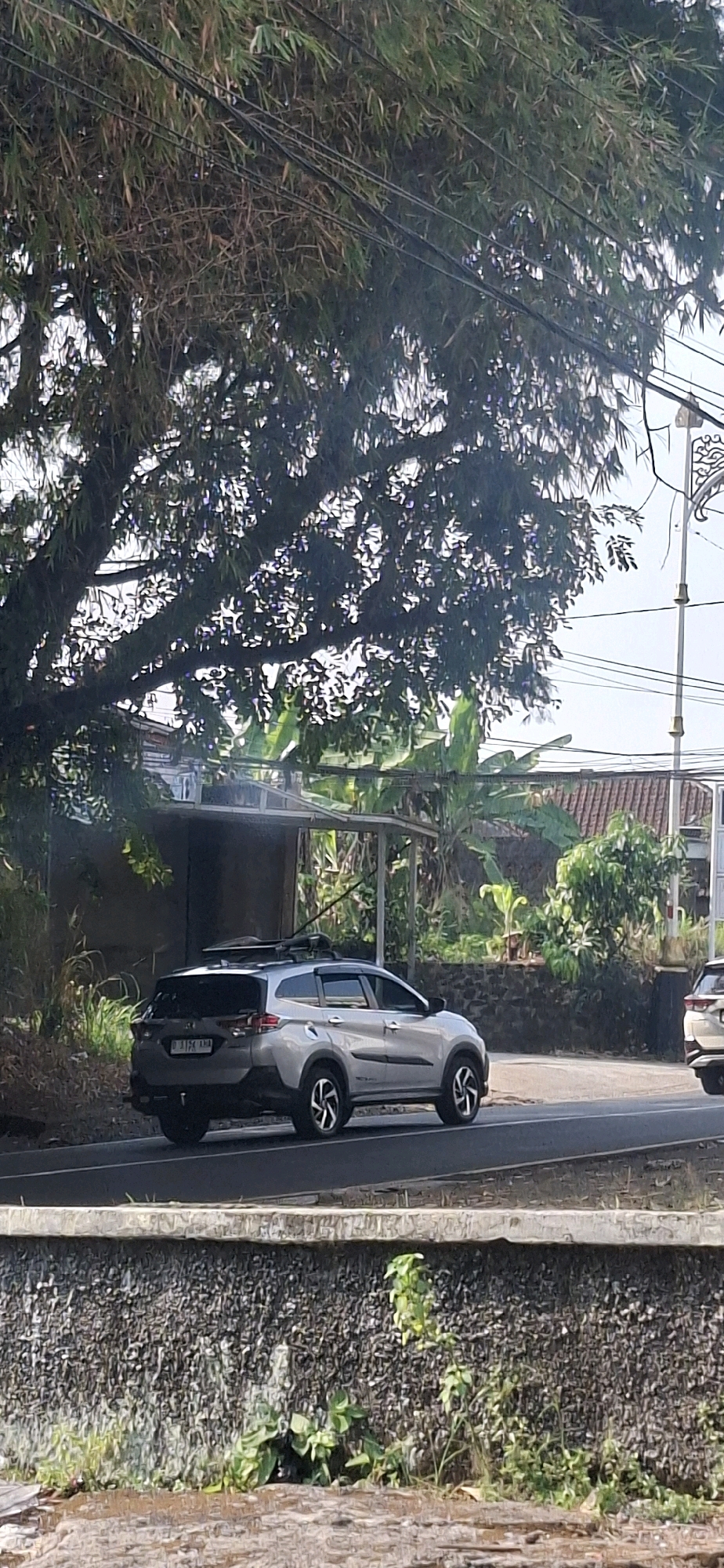 A silver SUV is parked on the side of a road in Cilaku, Indonesia. The car is a Toyota Rush. The SUV has a roof rack and is facing to the right of the image. In the background, there are trees and a building. The road appears to be paved, and there is a concrete barrier on the left side of the image. The sun is shining, casting shadows on the road. There are power lines running overhead.