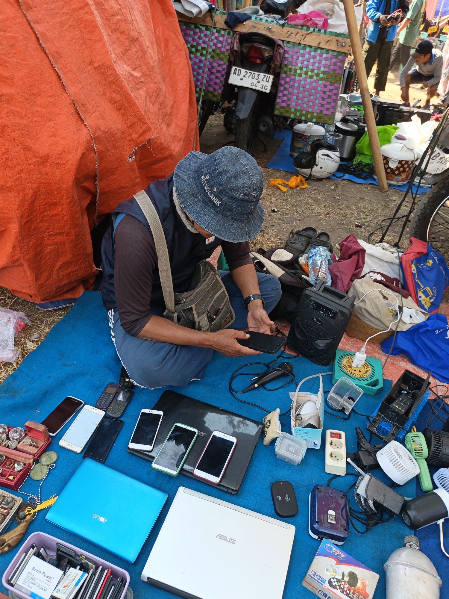 In Jatinom, Indonesia, on a bright day, a man wearing a blue denim hat and a dark shirt with a light-colored strap bag is seated on a blue tarp on the ground. He is holding a black smartphone in his hands. The tarp is laid out in what appears to be an outdoor market setting, with various items spread around him.
To his left, a large, wrinkled orange tarp creates a backdrop. A collection of jewelry, including rings and necklaces, are displayed in red and brown open boxes. Several round, flat stones and a beaded necklace are also present.
In front of him, a collection of electronics is laid out on the blue tarp. These include multiple mobile phones of various colors (white, black, green, and red), a blue ASUS laptop, and a white ASUS laptop. There is also a pink container filled with batteries and small electronic components.
To his right, a black portable speaker with a mesh front and some controls is visible. Wires and a microphone are connected to it. Several other electronic accessories are scattered around, including chargers, a white power strip with three outlets, and a small green desk fan. Other items present include a black power bank, a dark blue and white object, and a metal water bottle.
In the background, a motorcycle with a license plate that reads "AD 2703 ZU" is parked. The motorcycle is decorated with a colorful woven panel in shades of pink and green. Several helmets, including a white and black one and a white one with a pattern, are placed on the ground near the motorcycle. Other people are visible in the background, appearing to be customers or vendors at the market. The ground surface is a mix of dirt and scattered debris.