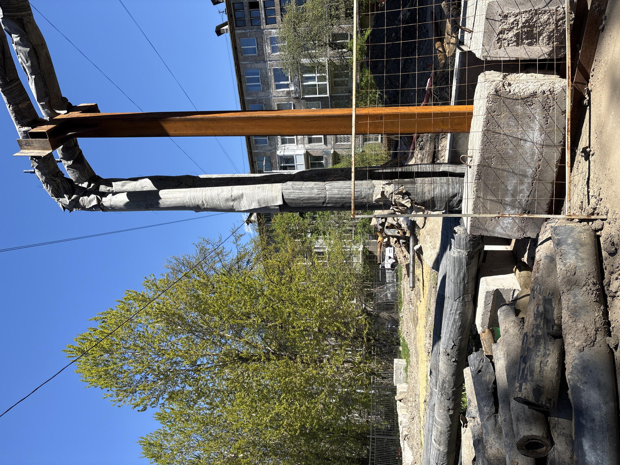 The image depicts a construction or renovation site, likely in Saint Petersburg, Russia, given the architectural style of the buildings in the background. An exposed network of insulated pipes supported by a steel beam is visible. Concrete blocks, some reinforced with rebar, are clustered near a wire fence. A pile of large, possibly industrial, pipes lie on the ground. In the background, a multi-story apartment building with numerous windows rises. A tree with light green leaves fills the left foreground. The sky is clear and blue. No people are visible in the immediate scene.