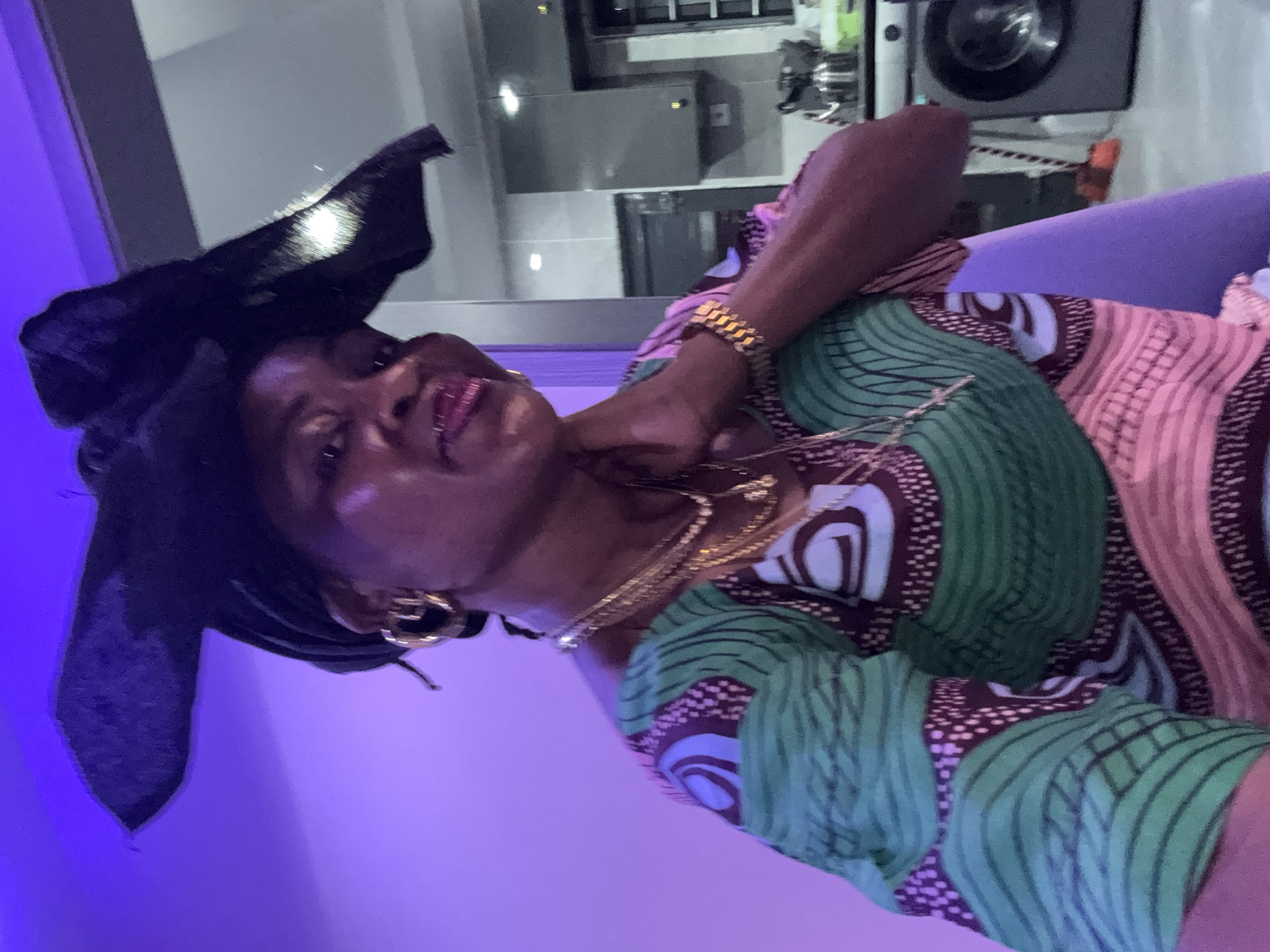 In Ajara, Nigeria, a woman is captured in a selfie, her face illuminated by purple light. She is adorned with a black headwrap, large gold hoop earrings, and multiple gold necklaces. Her off-the-shoulder dress features a vibrant pattern of green, pink, and maroon. Her left hand rests on her neck, and a gold watch is visible on her right wrist. Behind her, a domestic scene unfolds: a gray kitchen cabinet with a white backsplash is visible, along with a washing machine and a broom leaning against the wall. The background lighting shifts from the dominant purple to a softer, neutral tone, suggesting an indoor environment.