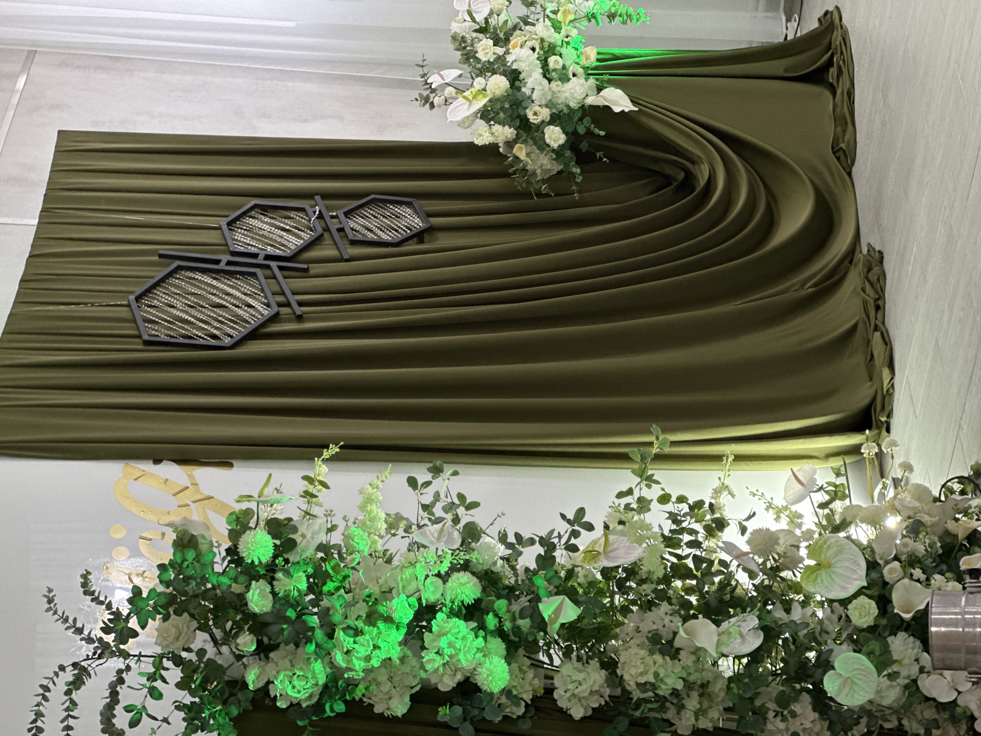 The image shows a decorated wall, likely at an event venue in an unknown city. The wall is draped with a heavy, olive-green fabric arranged in sweeping curves and folds. Three decorative geometric figures, resembling connected hexagons filled with textured material, are attached to the fabric. A floral arrangement, consisting of white and green flowers, is positioned on the upper right of the draped fabric. Below, a larger arrangement with more flowers and foliage is situated, with some of the blooms illuminated by green lighting. A gold-colored inscription or design is visible on the wall behind the lower floral arrangement. The overall scene suggests a formal or celebratory setting.