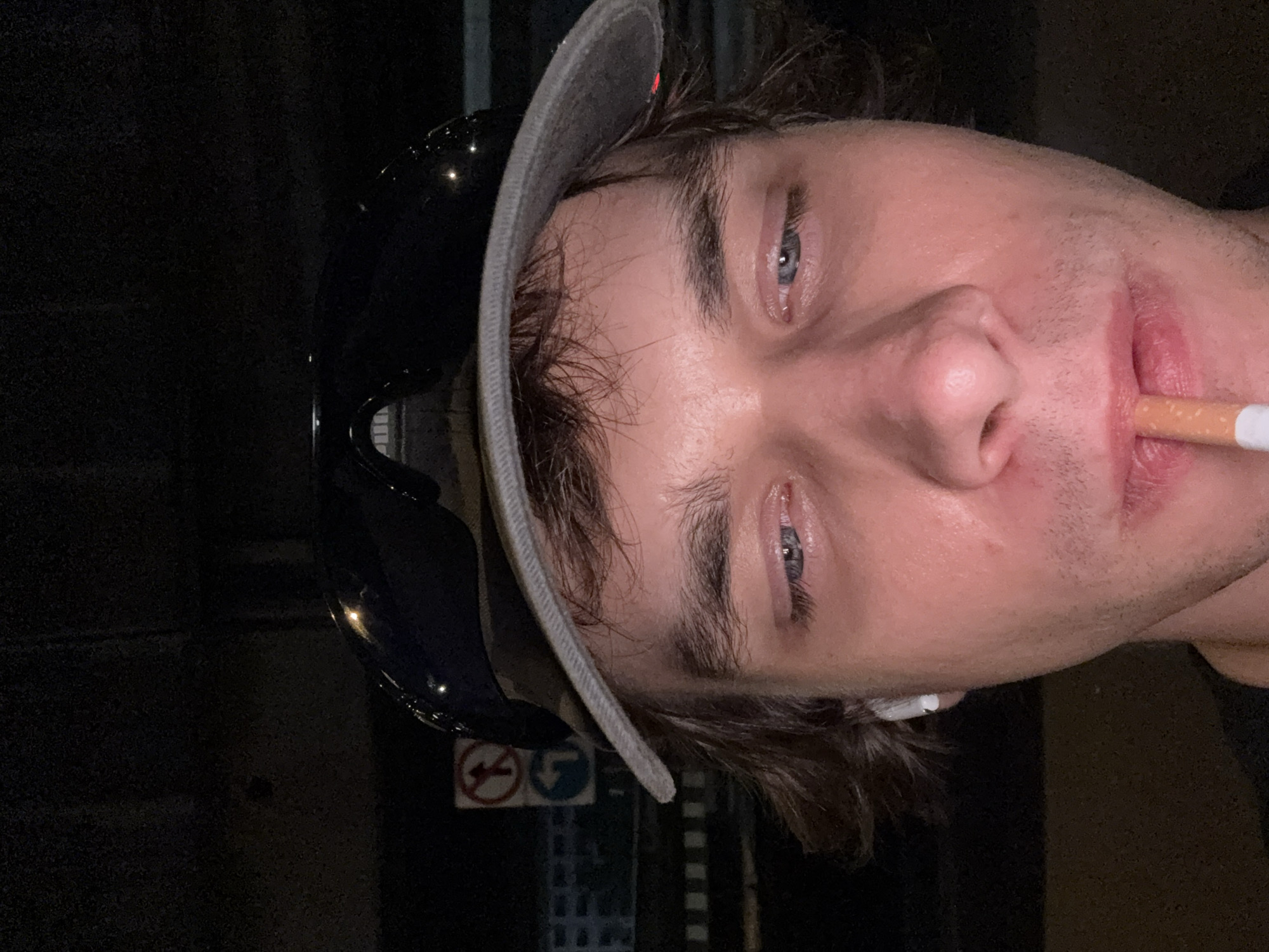 A close-up portrait of a fair-skinned individual with light blue eyes, wearing a grey baseball cap and dark-rimmed glasses atop the cap. The person has a cigarette held between their lips. The background is dark, appearing to be nighttime, with a glimpse of a street sign indicating traffic directions (no U-turn, turn left) and hints of urban infrastructure, potentially placing the scene in a city environment. The setting is consistent with a street or public space. Bangkok, Thailand, cannot be verified from this image.