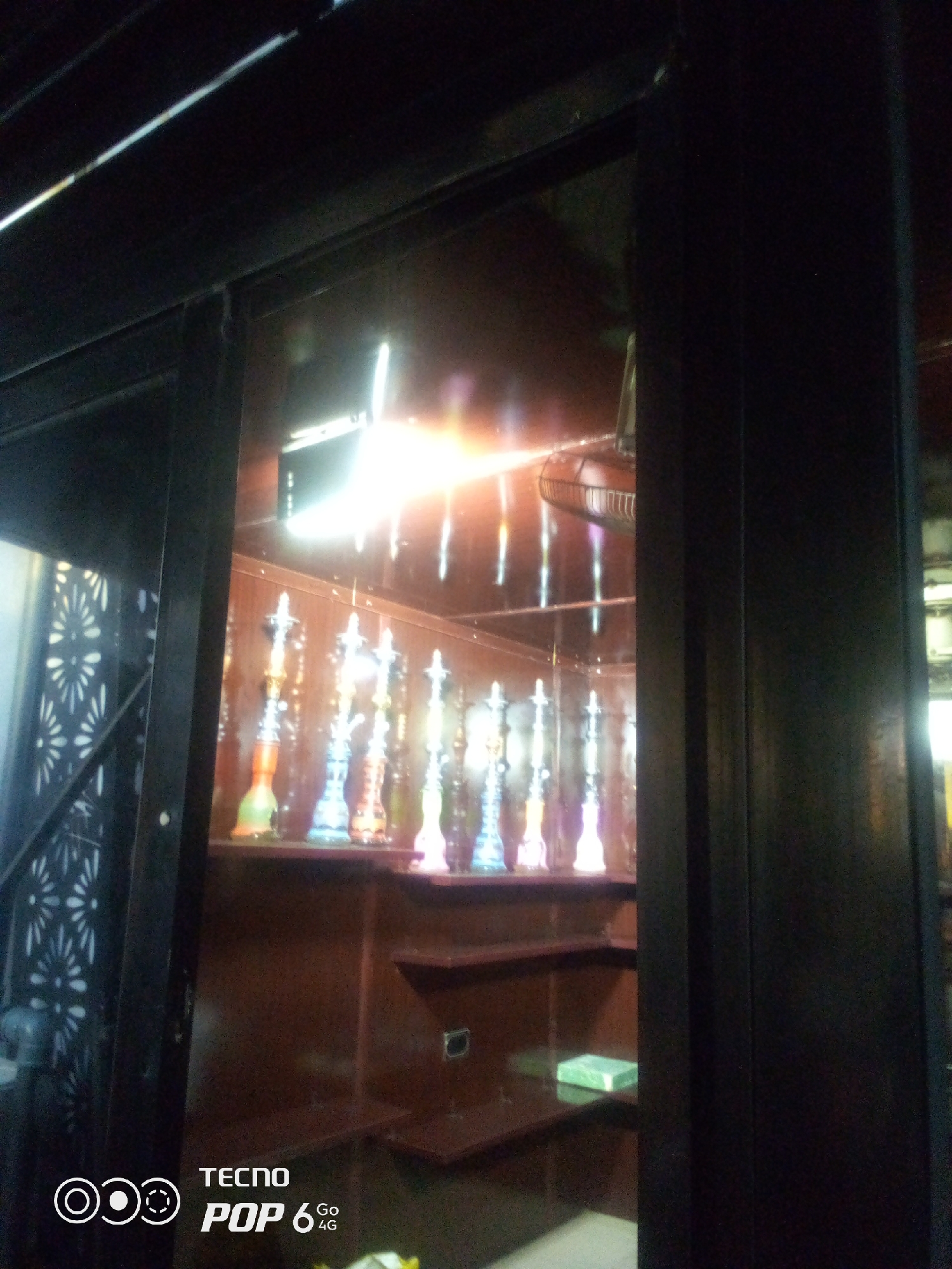 The image shows a display case with multiple colorful shisha pipes arranged on shelves. The pipes vary in color, including shades of green, blue, orange, and purple. The display case has a dark wooden finish, and the background is reflective, showing lights and the ceiling. To the left of the display case, there is a patterned screen. In the lower left corner, a Tecno Pop 6 4G logo is visible. The scene appears to be an indoor establishment, possibly a shop or lounge, located in Howayi, Nigeria. There are no people visible in the image.