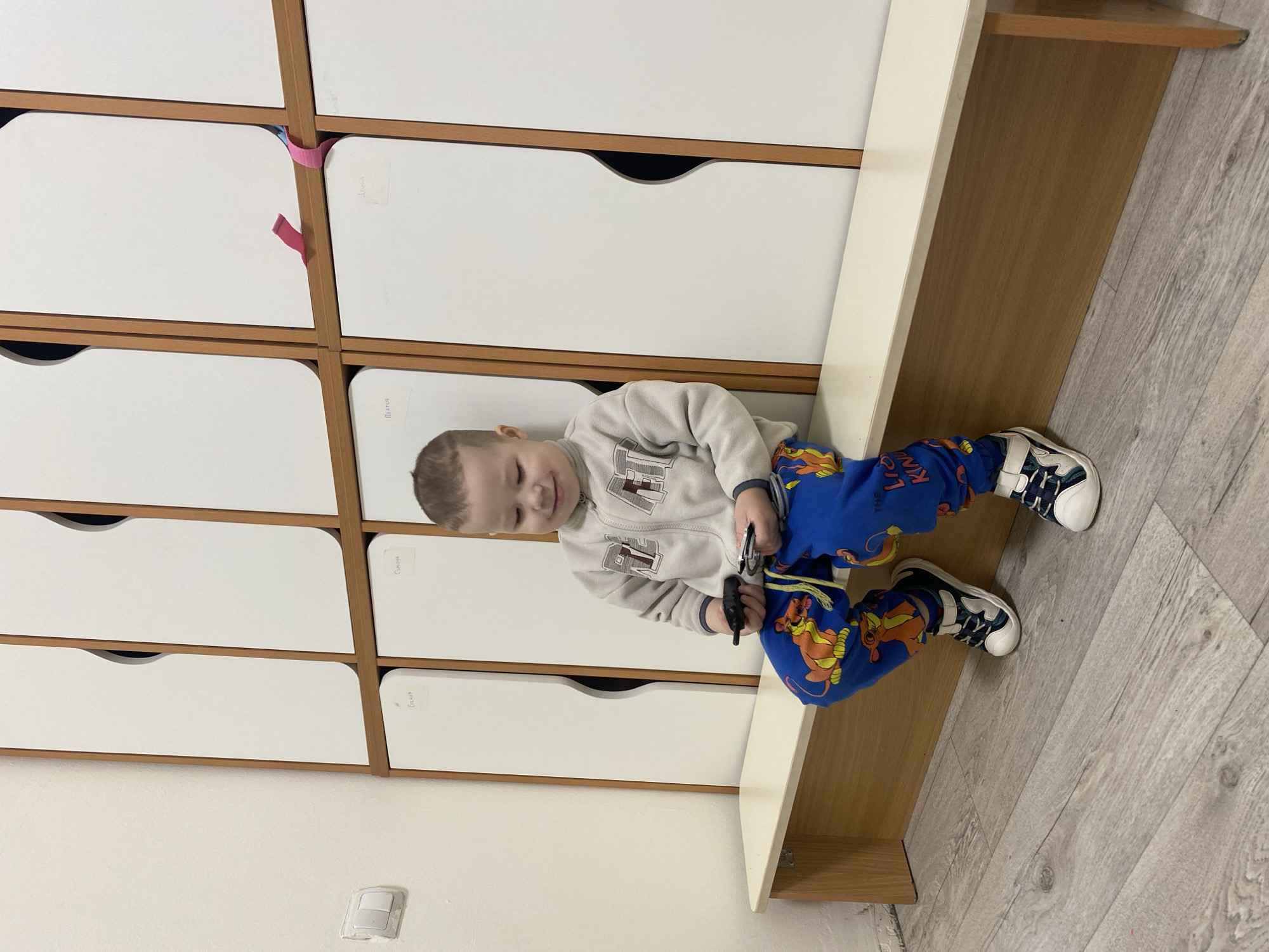 A young boy is sitting on a wooden bench against a wall of white storage lockers. He is wearing a gray sweatshirt, blue pants with "The Lion King" print, and white sneakers with black and blue accents. He is holding a black object in his hands. The floor is gray, wood-like. A light switch is on the wall. The location cannot be determined as Sochi, Russia, based on the visible elements in the image.