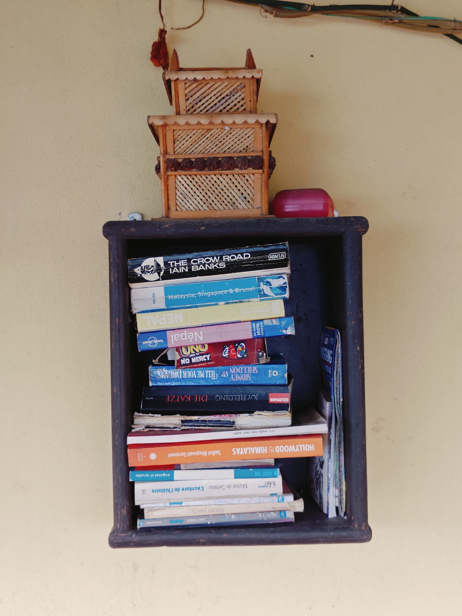 The image shows a shelf with a stack of books and two wooden decorative boxes. The shelf is dark wood and appears to be mounted on a light yellow wall. The decorative boxes are on top of the shelf, with the smaller one on top of the larger one, and are made of wood with a lattice design. A red, oval-shaped object is placed next to the boxes on the shelf.
The books are stacked vertically in the shelf, with titles visible on their spines. The visible titles include "THE CROW ROAD" by Iain Banks, "Malaysia, Singapore & Brunei" (Rough Guides), "NEPAL", "Nepal" (Lonely Planet), "UNO NO MERCY", "TELL ME YOUR DREAMS" by Sidney Sheldon, "DIE KATZE", "HOLLYWOOD HIMALAYAS", "L'écriture de l'histoire", and "SACRED VINYASA" by ROYINA GREWAL. There are also several other books with less visible titles. A folded magazine or pamphlet with the title "LANGTANG" is also visible in the stack.
The scene suggests a personal collection of books and decorative items, possibly in a home or a small shop. The presence of books about Nepal and travel guides to the region, along with the wooden decorative boxes, might indicate a connection to Nepal or a specific interest in its culture and geography.
The image does not provide explicit information about the specific location within the city of Pokhara, Nepal. However, the style of the decorative boxes and the type of books could be indicative of a traditional or cultural setting in Nepal. The overall environment appears to be indoors or in a covered outdoor space, given the absence of natural light variations and the presence of a wall.