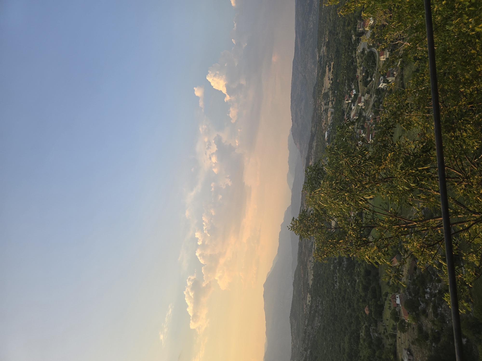 This is a high-angle, wide shot photograph of a rural landscape at sunset. In the foreground, a dark metal railing runs horizontally across the bottom of the frame. Behind the railing, the branches and leaves of several trees dominate the lower half of the image, casting shadows and partially obscuring the view.

The middle ground reveals a valley with scattered houses, predominantly featuring red-tiled roofs. A winding road is visible, connecting the houses. The valley floor is a mosaic of green trees and patches of lighter vegetation. In the distance, a series of rolling hills and mountains stretch towards the horizon, their forms softened by the atmospheric haze and the fading light.

The sky above is a gradient of soft blues and warm, peachy-orange hues, indicating sunset. Scattered white and yellow clouds are illuminated by the setting sun, adding texture and depth to the sky. The overall impression is one of serene, rural beauty bathed in the warm glow of twilight.

The location is identified as Gornja Vrbica, Montenegro.
