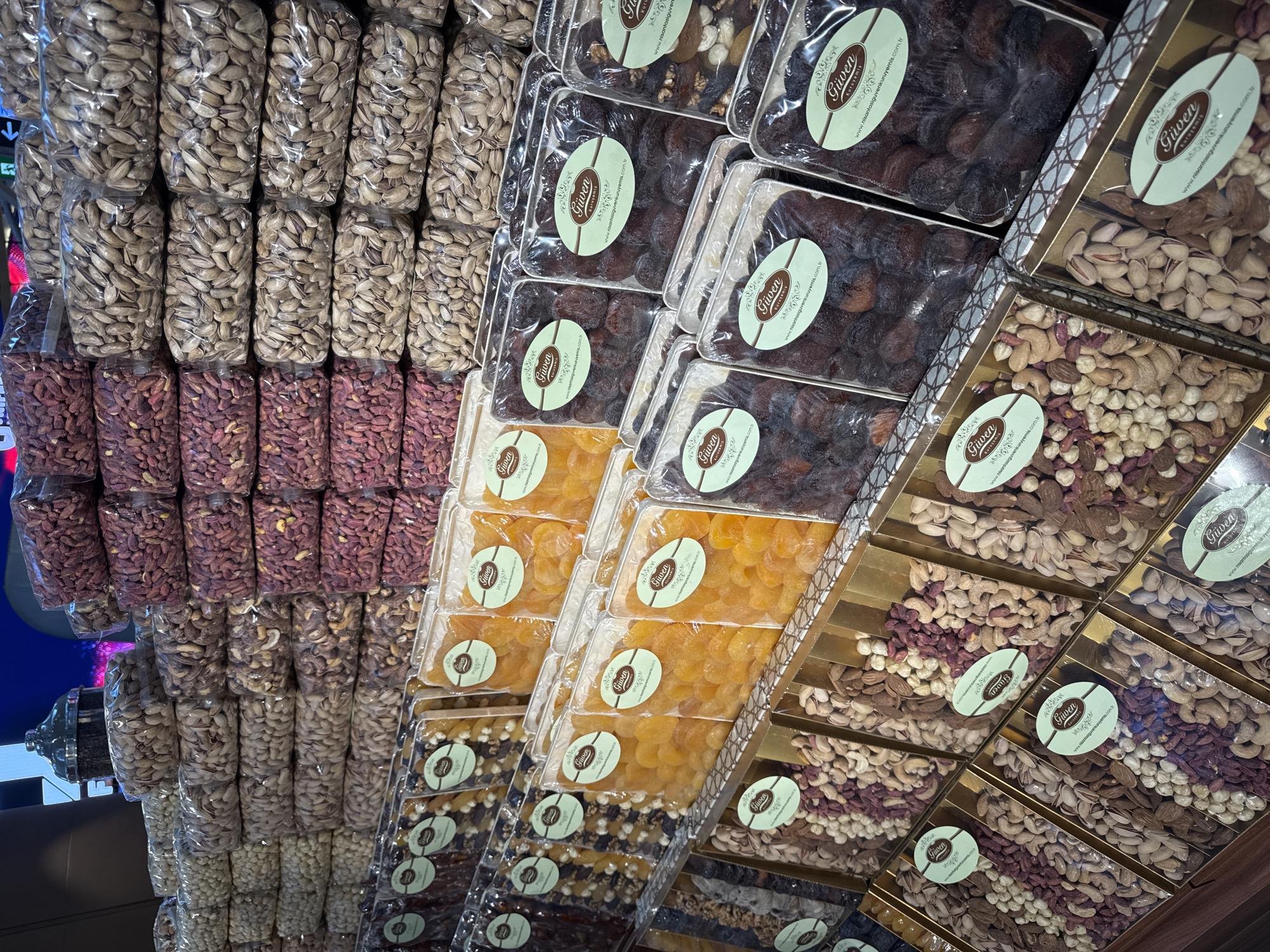 This image captures a vibrant and densely packed display of packaged dried fruits and nuts, likely in a specialty food store or market in Arnavutköy, Turkey. The scene is an interior retail setting, illuminated by artificial light, with products meticulously arranged on shelves.
The primary subjects are a wide assortment of nuts and dried fruits, presented in various types of packaging. On the left side, tall stacks of clear plastic bags are visible, filled with pistachios in shells (light green/beige), and below them, bags containing reddish-brown, elongated nuts, possibly a type of roasted peanut or flavored pistachio kernels.
Towards the center and right, an array of clear plastic containers and ornate gold-colored gift boxes showcases dried apricots in two main varieties: dark reddish-brown ones and bright orange/yellow ones. These containers are neatly stacked, creating an organized visual pattern. The gold boxes are particularly notable, filled with meticulously arranged assortments of mixed nuts, including pistachios, cashews, and almonds, often separated into sections.
Each package and box features an oval-shaped, light green label bearing the brand name "Güven Kuruyemis." The labels also display the website address "www.nisantasiguvenkuruyemis.com.tr", indicating the origin or main branch of this business, likely referring to Nişantaşı, a district in Istanbul, where "Güven Kuruyemis" is a known establishment.
No people are visible, and there is no activity taking place other than the static display of goods for sale. The overall impression is one of abundance and quality, with the products presented attractively to potential customers. The neat arrangement and uniform branding suggest a well-managed and professional retail environment.