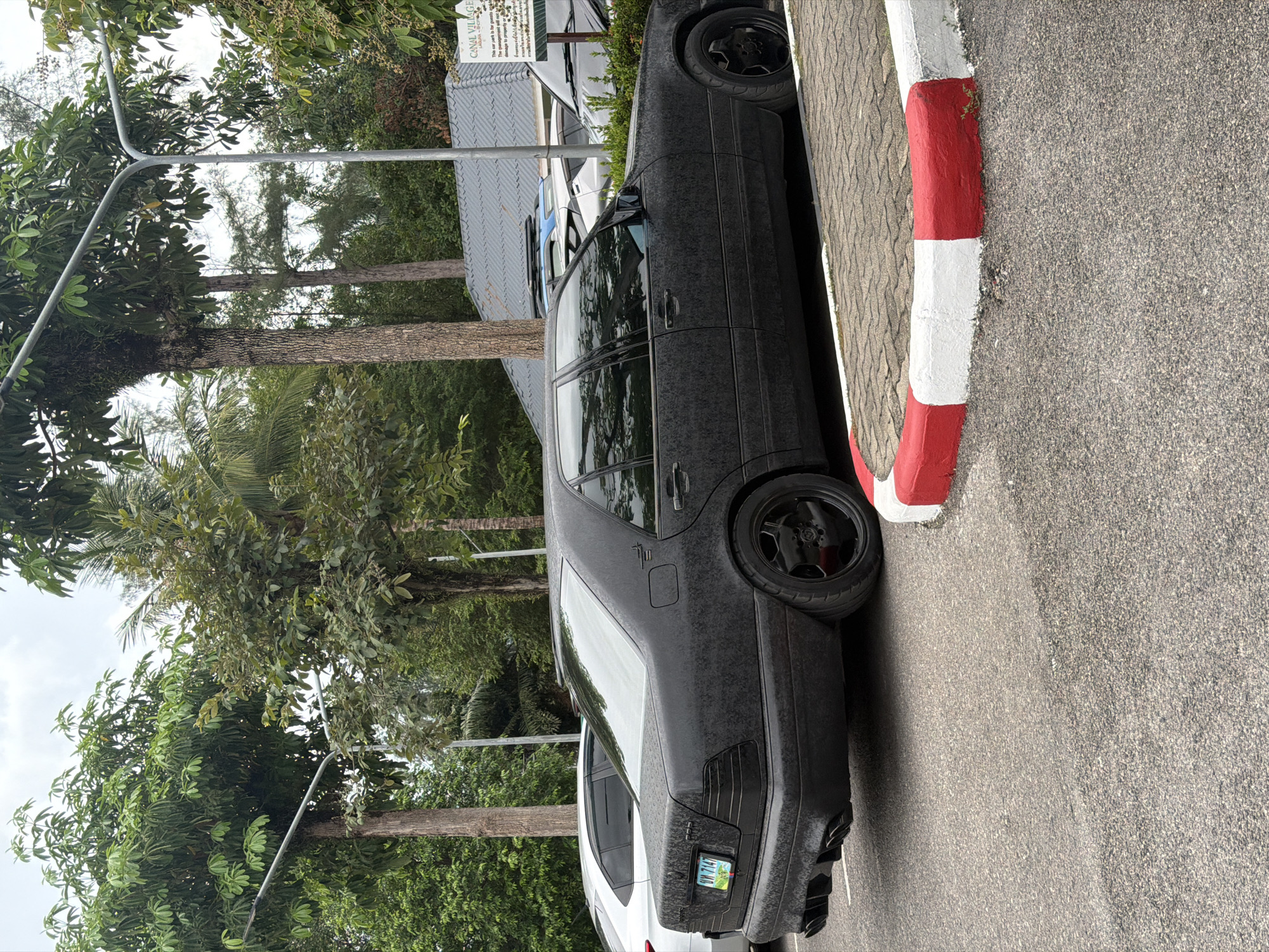 A matte black Mercedes-Benz S-Class sedan is parked in a lot with a red and white painted curb in the foreground. The car has a textured finish and is seen from the rear passenger side. Several trees are visible in the background, along with a gray-roofed building with a sign that reads "CANAL VILLAGE". The ground is paved with asphalt and cobblestones. The sky is overcast. This scene is located in Choeng Thale, Thailand.