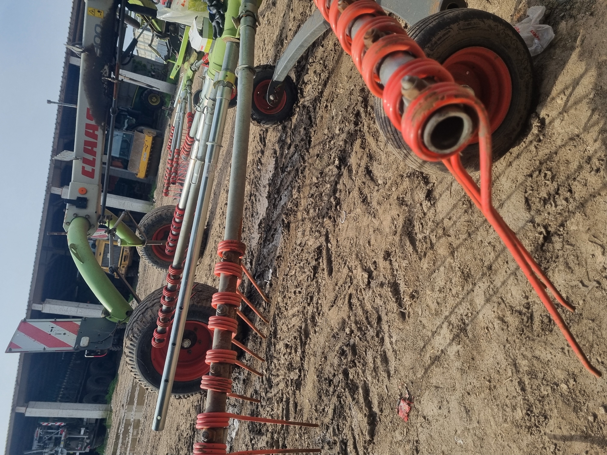 The image shows a hay rake implement on muddy ground in Ovcharovo, Bulgaria. The rake features multiple red tines attached to a metal frame with small wheels. A Claas tractor is partially visible in the background. The ground is covered in dirt and tire tracks.
