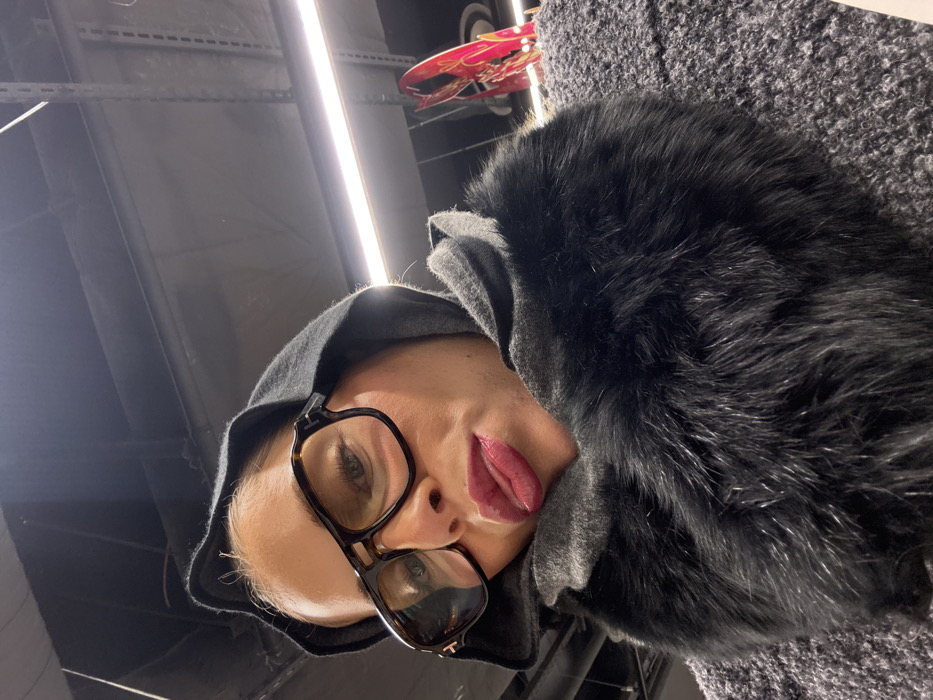 The image is a low-angle, close-up selfie of an adult woman taken indoors. She is looking directly at the camera with a playful expression, her tongue slightly protruding from her dark red-lipsticked mouth. She wears large, tortoise-shell framed eyeglasses, a dark hooded garment over her head, and a substantial dark grey or black faux fur collar. Her outer garment appears to be a textured grey coat. The background consists of a dark ceiling with exposed utilities and a bright, horizontal LED light fixture. A partial red and pink decorative object is visible on the right side of the frame, above and behind her right shoulder. The specific location within Paris, France, is not discernible from the image content.