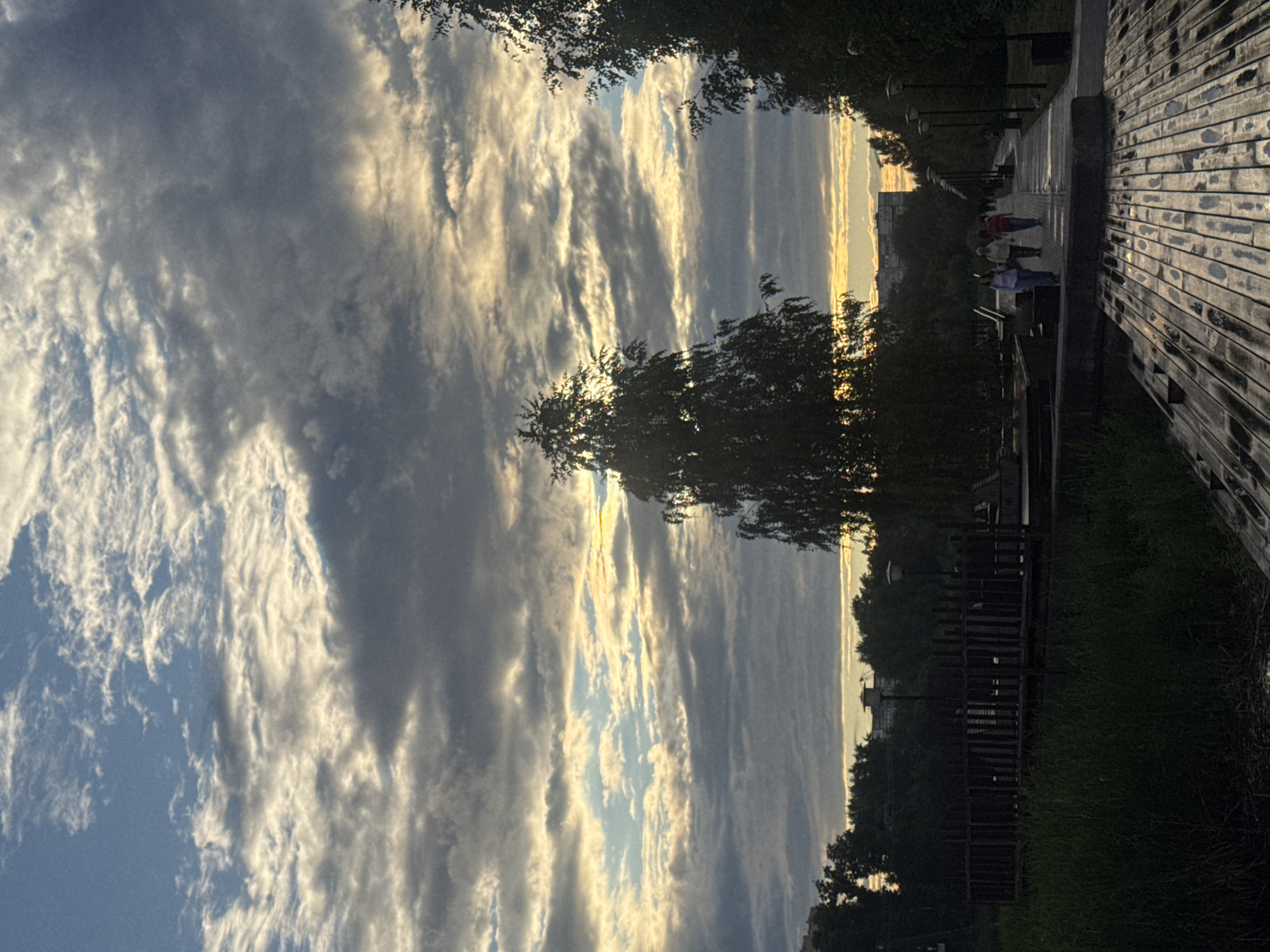 This image depicts a sunset scene along a wooden walkway. The sky is filled with dramatic, elongated clouds catching the warm hues of the setting sun, creating a striking contrast between light and shadow. The sun itself is low on the horizon, casting a golden glow that illuminates the undersides of the clouds.
The walkway, constructed of weathered wooden planks, runs along the right side of the frame, appearing wet, possibly from recent rain or dew. On the left side of the walkway, a line of trees and lampposts suggests a park or recreational area. In the distance, silhouetted figures of people can be seen walking along the path, their forms indistinct against the backdrop of the sky and trees. Further beyond, buildings are faintly visible, indicating an urban or suburban setting bordering the natural landscape.
The overall mood is serene and contemplative, enhanced by the fading light and the stillness of the scene. The time of day is clearly sunset. The weather appears to be clear with cloud cover. The location is suggested to be Nizhny Novgorod, Russia, by the provided context. No visible text is present in the image.