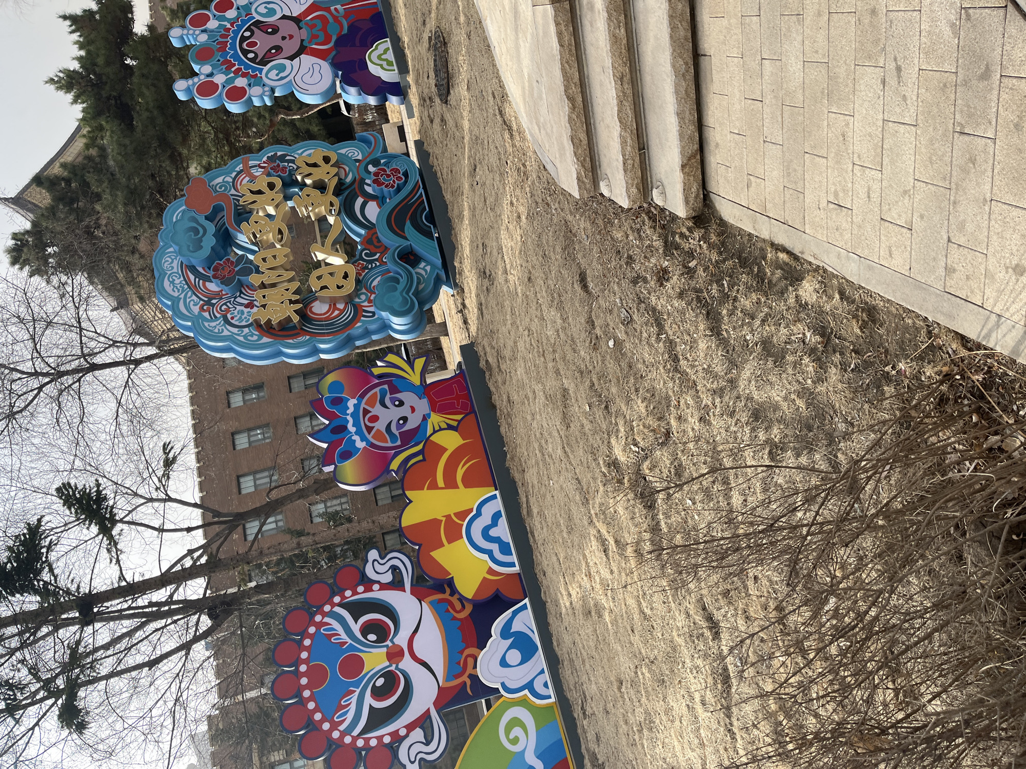 The image shows a row of decorative signs placed along a walkway or park path. The signs feature colorful, stylized designs that appear to be based on traditional Chinese opera masks or imagery. One sign has gold colored characters that appear to be Chinese script. The background includes a bare tree, a brick building, a patch of dry grass or dirt, and some paving stones or steps. It is the city of Changchun City, China.