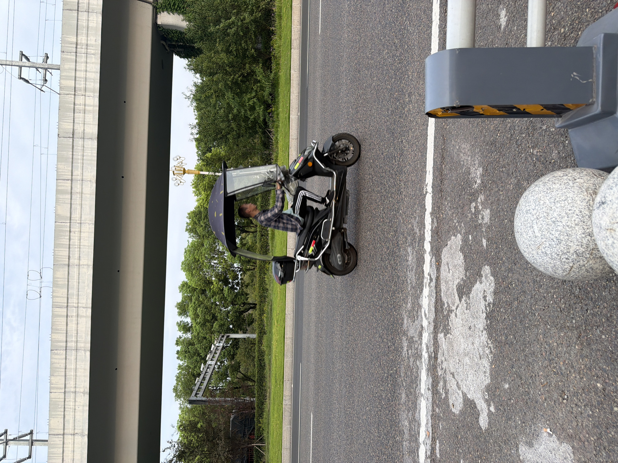 A person is riding a black electric scooter with a clear windshield and a dark umbrella overhead. The scooter has a black storage box mounted on the rear. The person is wearing a plaid shirt and dark pants. The scooter is on a paved road with a white line marking the lane. In the background, there are trees, a grassy median, and a concrete overpass. A street lamp with multiple lights is visible behind the scooter. The scene appears to be in an urban or suburban area of Jiangjin, China. The lighting suggests it is daytime. There are no other people visible in the immediate vicinity of the scooter.