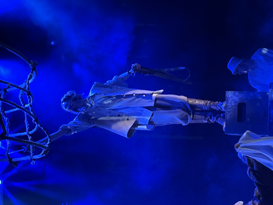 This image depicts a theatrical performance or stage event under intense blue lighting in Novoshakhtinsk, Russia. A central figure, likely an actor, stands elevated on a dark, rectangular stage block. This individual is blindfolded, wearing a light-colored layered costume consisting of a shirt, an open vest with grommets, loose trousers, and dark tall boots. Their right arm is raised, grasping a dark, coarse rope net suspended above, while their left hand holds a dark, loop-ended strap. Partially visible in the lower frame are two other individuals: one on the left in dark clothing with a white cloth, and another on the right in a light-colored top and bucket hat, observing the central figure. The environment is characterized by strong blue stage lighting and visible atmospheric haze.