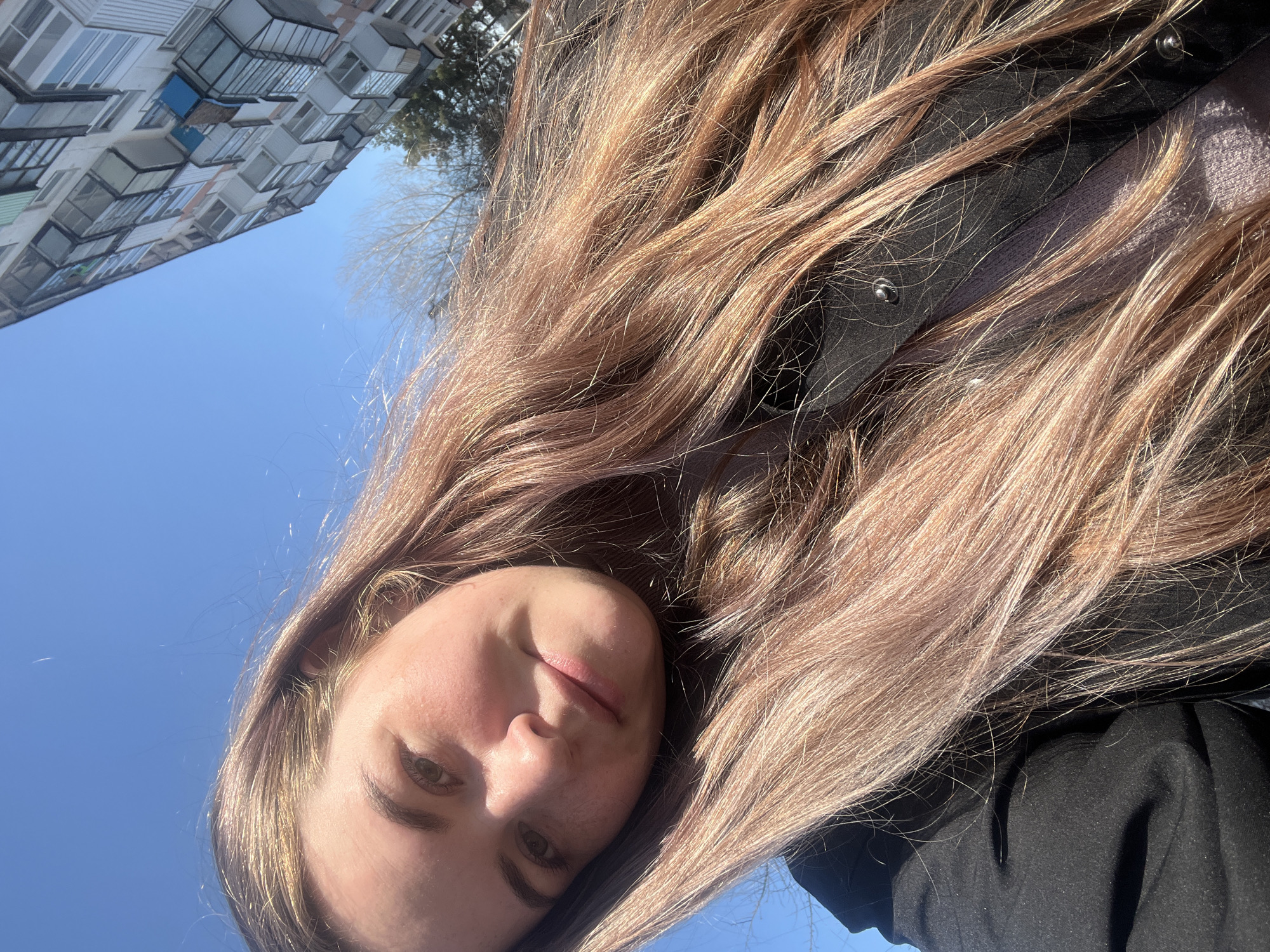 A photograph depicts a light-skinned person with long, brown hair outdoors, likely in Chaikovsky, Russia. The person's face is angled toward the lower left corner of the image, while a section of a multi-story building is visible in the upper left corner against a clear blue sky. The person is wearing a black coat. Bare tree branches are also visible in the upper portion of the image.