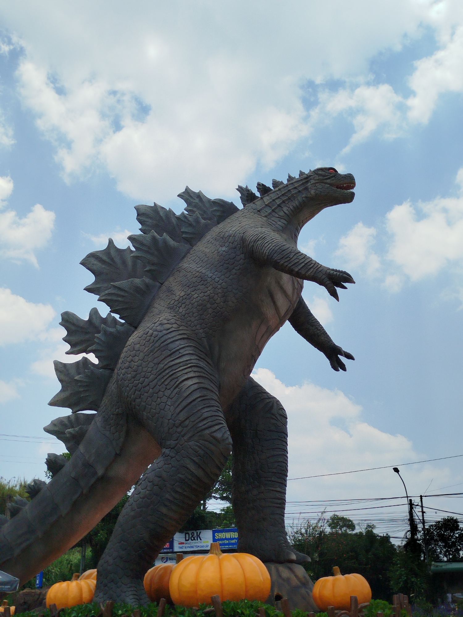 A photograph captures a Godzilla statue surrounded by pumpkins under a partly cloudy sky, likely taken in Cam Ly, Đà Lạt, Vietnam. The statue is a dark gray, with a scaly texture and dorsal fins. It appears to be standing on a platform amid a collection of bright orange pumpkins. In the background, there are signs, possibly related to a showroom, along with trees and shrubbery. The statue and pumpkins are bordered by a low wooden fence. Power lines and a street lamp are visible in the distance. The overall scene seems to combine the iconic figure of Godzilla with an autumn or Halloween-themed display.