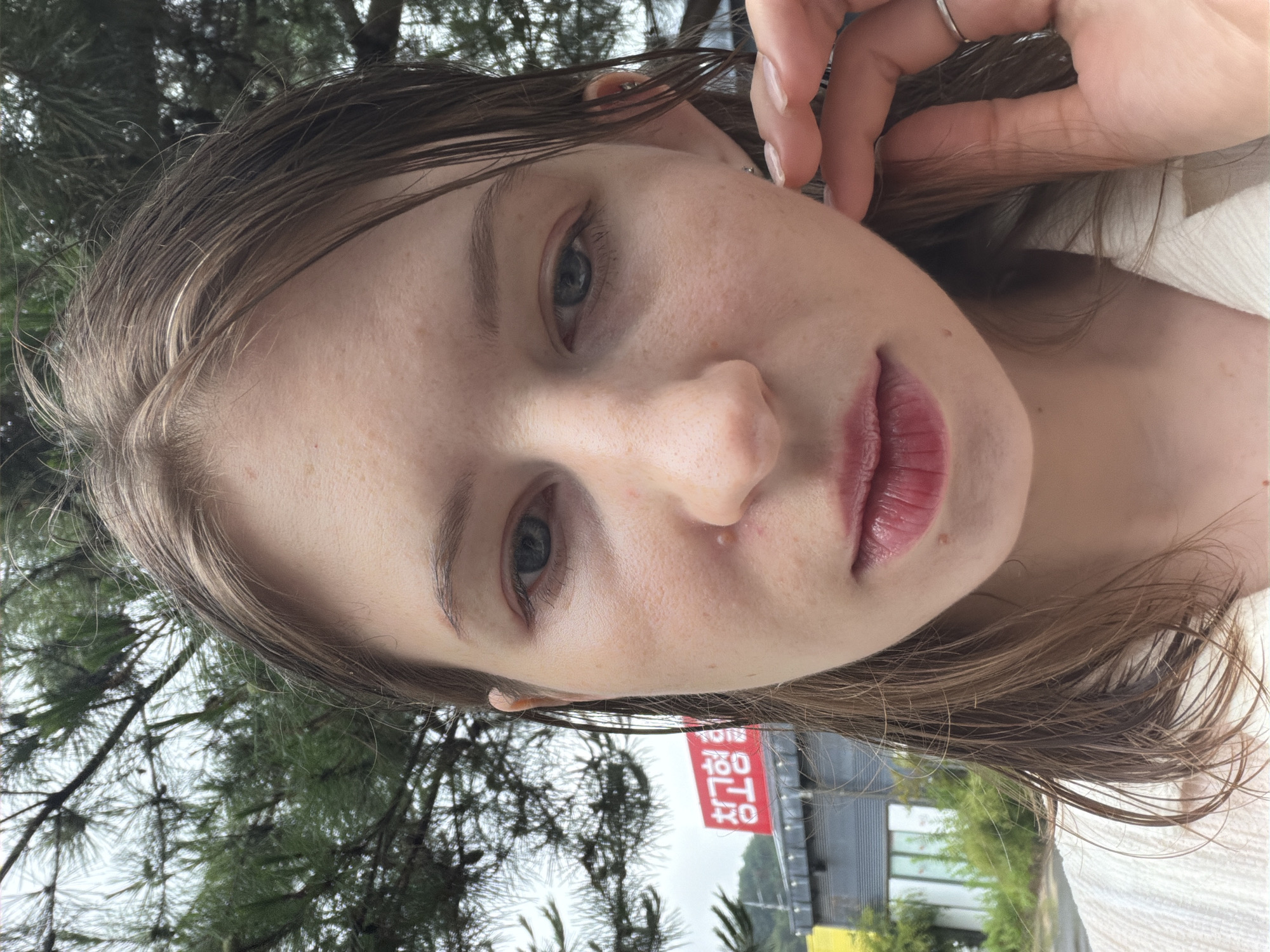A close-up, frontal, outdoor, medium shot shows a young Caucasian woman with light brown hair, which appears wet, partially obscuring her face. Her skin is fair with light freckling, and she has blue eyes. She is looking directly at the camera with a neutral expression. Her lips are parted, revealing a hint of teeth, and are glossy with a reddish hue. A small mole is visible below her lower lip. Her right hand is raised, touching her right ear and cheek, with her thumb and index finger gently pinching her earlobe. She is wearing a light-colored, textured top and a silver ring on her right ring finger.
In the background, a red and white sign with Korean characters (청 고 형) is visible to the left of the woman's face. Behind the sign, a grey, low-rise building with windows can be seen. Pine trees with green needles frame the upper portion of the image, their branches extending over the woman's head and hair. The sky is overcast and appears grey. The overall setting suggests a slightly damp or humid environment. The location is identified as Pocheon-si, South Korea.