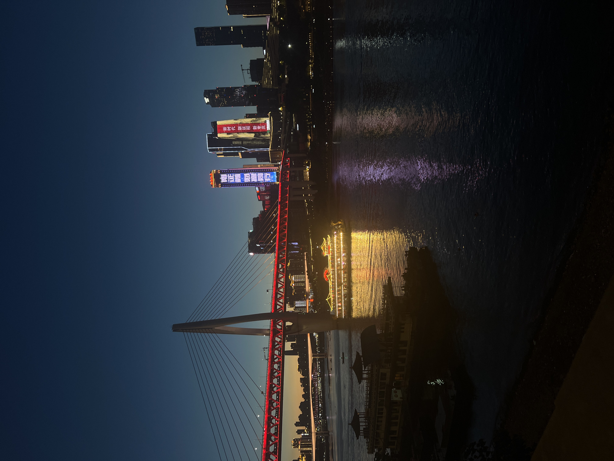 This is a wide-angle, horizontal shot of the Yuzhong District in Chongqing, China, at dusk. The scene is dominated by the Yangtze River, reflecting the illuminated cityscape and a large suspension bridge. The bridge, adorned with vibrant red lights along its structure, spans across the river. On the river, two traditional-style tour boats are visible. The boat in the foreground is dark and silhouetted, while the boat further back is brightly lit with golden lights, featuring ornate rooftops. In the background, a cluster of modern skyscrapers pierce the twilight sky. Several of these buildings are illuminated with colorful digital displays. The sky is a deep, clear blue, transitioning to a lighter hue near the horizon. The overall impression is one of urban grandeur and nighttime illumination.
