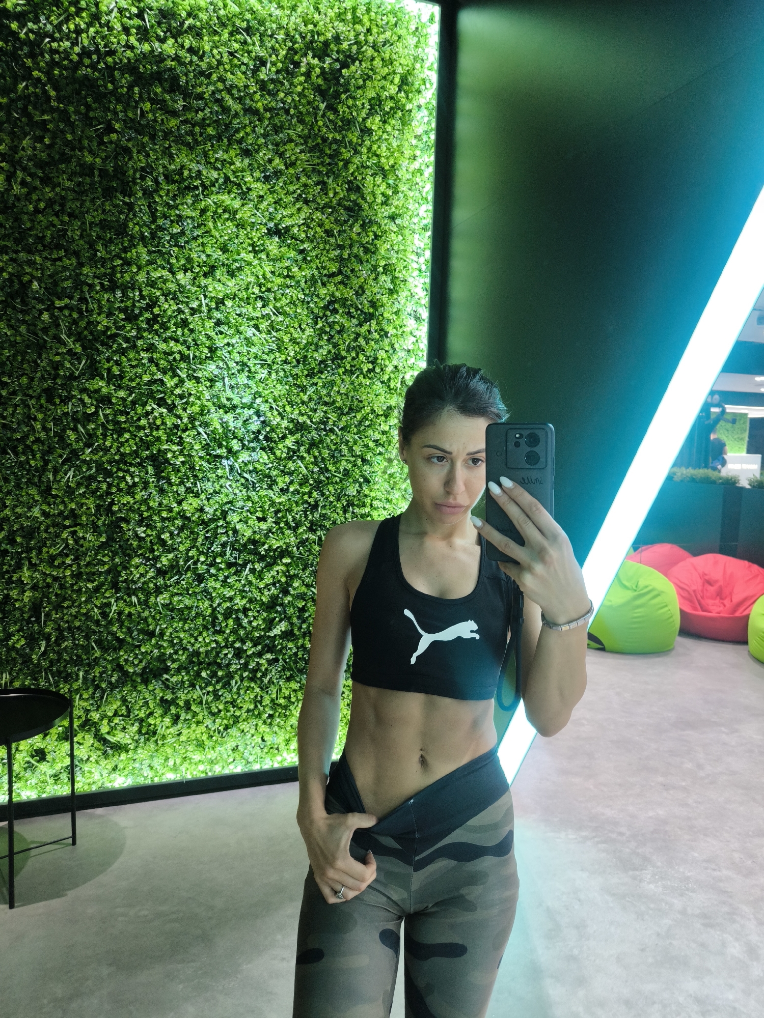 A person is standing indoors in front of a wall that has a panel of green foliage to the left and a black panel to the right with a linear blue light. The person is taking a selfie with a black smartphone, which is held in the right hand. They are wearing a black Puma sports bra and camouflage-patterned leggings. The left hand is holding the waistband of the leggings. Behind the person, there are several beanbag chairs in pink and lime green. A small black table is visible to the left. The setting resembles a gym or fitness studio. The city and location are not specified.
