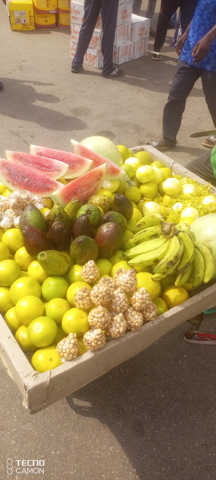 The image depicts an outdoor market scene in Jalingo, Nigeria, likely a roadside stall. In the foreground, a cart is filled with a variety of fresh fruits. Prominently displayed are several slices of watermelon, revealing their red flesh and white rind. Alongside the watermelon are numerous yellow lemons and green limes, creating a vibrant contrast. A bunch of unripe green bananas is also visible. Interspersed among the citrus fruits are several avocados, some appearing ripe and dark, others greener and firmer. Small, bagged portions of what appear to be nuts or seeds, possibly peanuts or groundnuts, are also present on the cart. In the background, blurred figures of people are visible, some standing, wearing casual clothing, and a stack of boxes, possibly related to merchandise, is also partially visible. The ground appears to be a paved surface, consistent with a street or market area. The lighting suggests it is daytime.