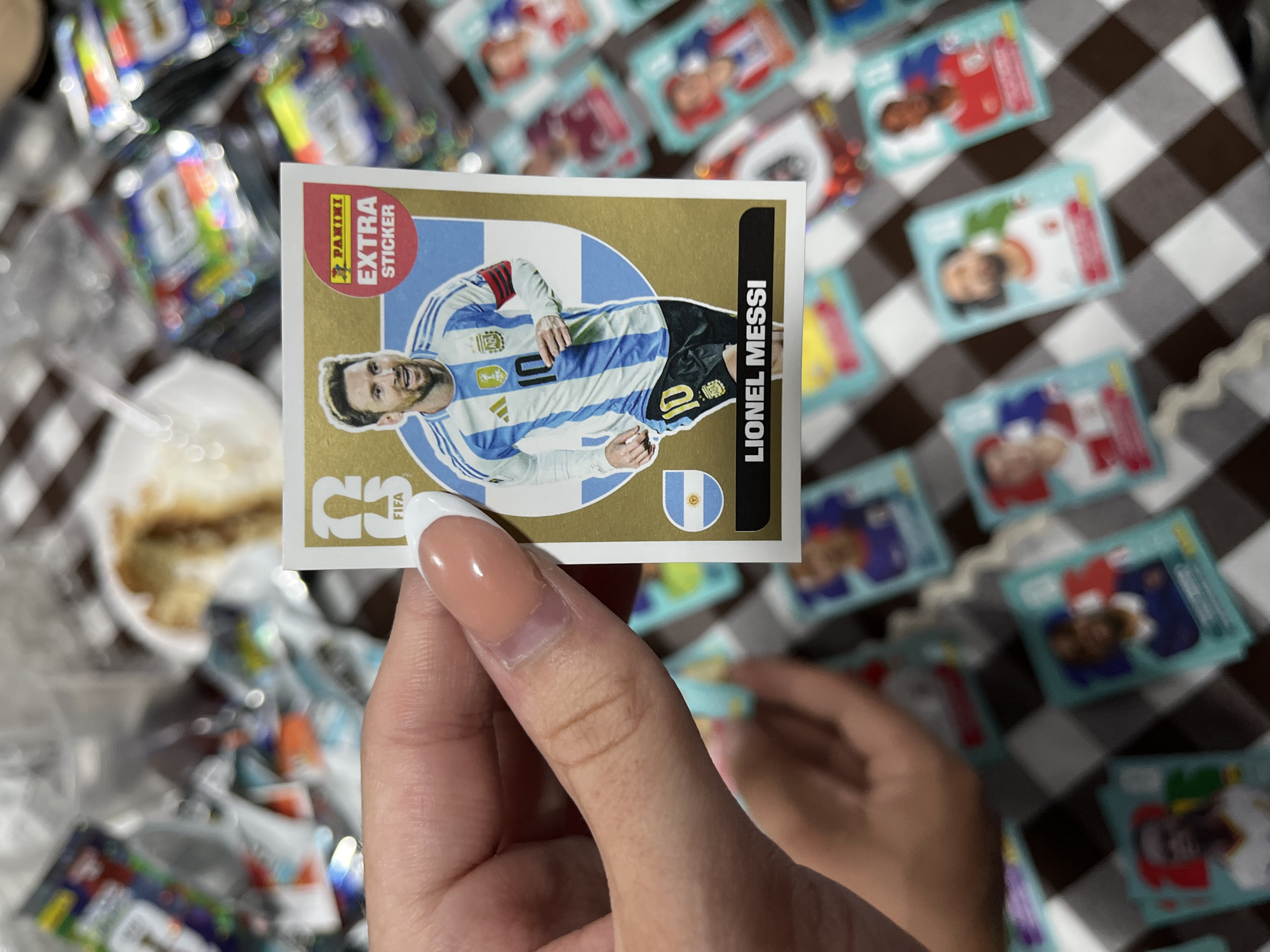 A close-up shot shows a hand holding a Panini FIFA World Cup Extra Sticker of Lionel Messi, against a background of other scattered FIFA World Cup stickers on a black and white checkered surface. The sticker features Messi in an Argentina jersey, set against a gold and blue design. Other stickers, wrappers and a plate with food are visible in the blurred background. The scene appears to be indoors. I do not have sufficient information to determine if this image was taken in San Bernardino Tlaxcalancingo, Mexico.