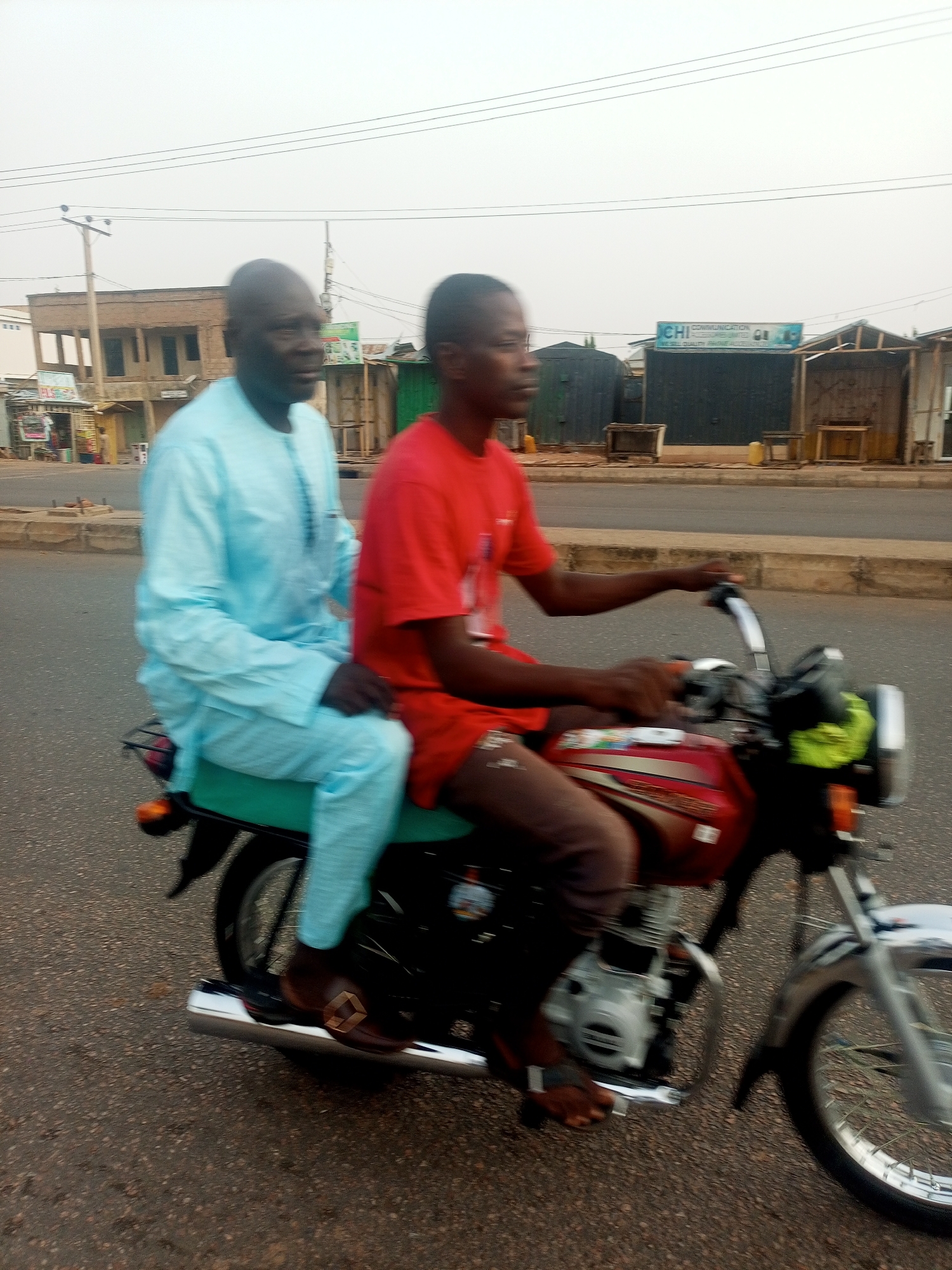 Two men are riding a red motorcycle on a paved road in Tempure, Nigeria. The man in front is wearing a red shirt and brown shorts. He is barefoot and driving the motorcycle. The man in the back is wearing a light blue traditional outfit and brown loafers. Buildings and shops are visible in the background.